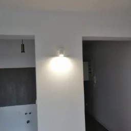 Wnętrze po odświeżeniu: białe ściany, punktowe oświetlenie LED, widoczne fragmenty podłogi i instalacji elektrycznej w dwóch przejściach.