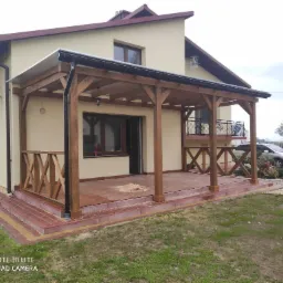 Drewniana konstrukcja zadaszenia tarasu przy domu jednorodzinnym, z widocznymi elementami balustrady w kształcie litery X, wykończona w ciepłym brązie, z fragmentami rozsypanego piasku na płytkach...