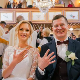 Uśmiechnięta para młoda w kościele prezentuje obrączki, na drugim planie goście i księża.