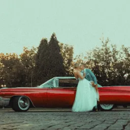 Pocałunek nowożeńców przy czerwonym, klasycznym Cadillacu Eldorado z białym dachem na tle drzew i nieba o zachodzie słońca.