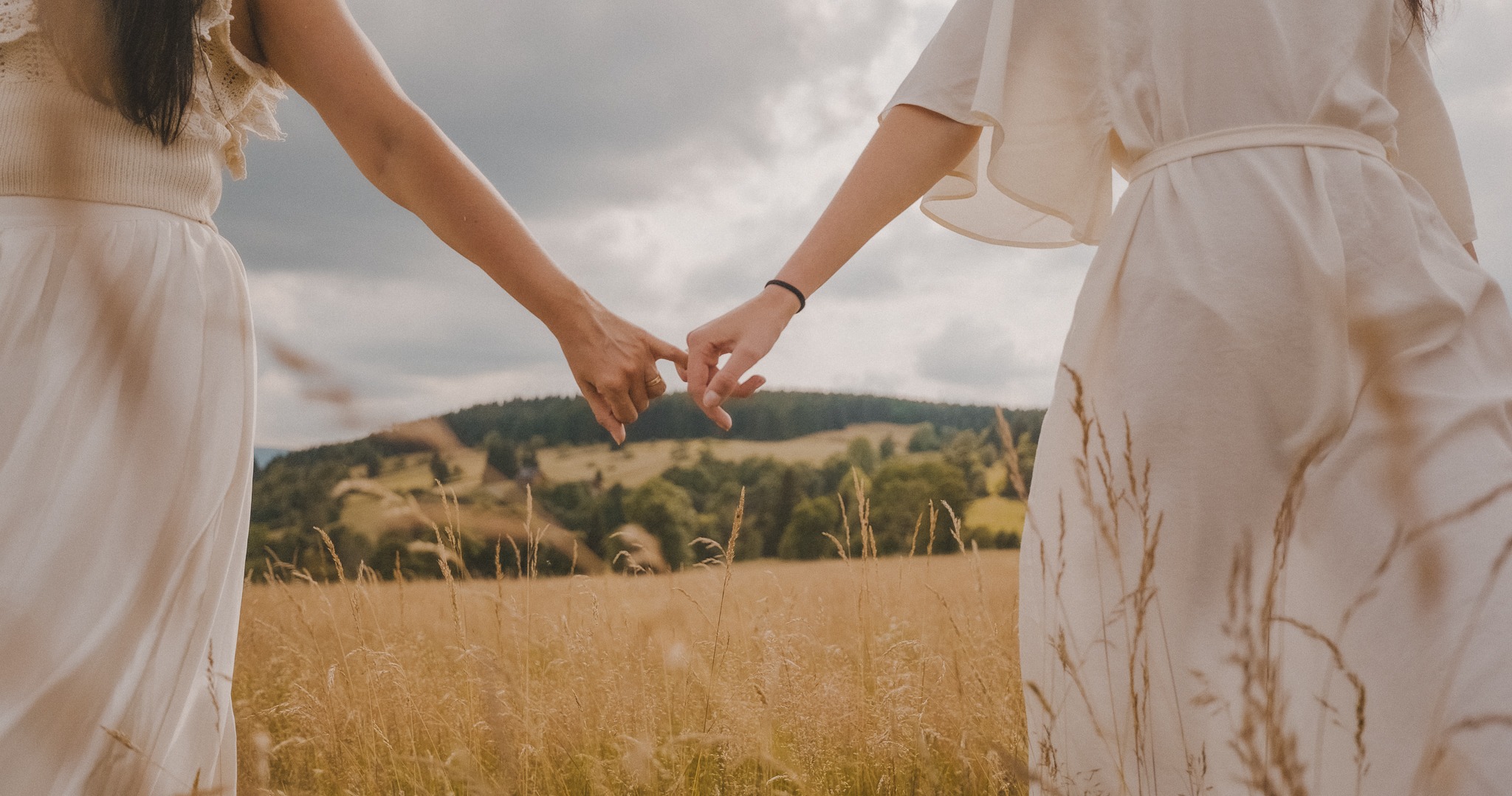 Dwie kobiety w zwiewnych, kremowych sukienkach trzymają się za małe palce na tle łąki i zalesionego wzgórza pod pochmurnym niebem.