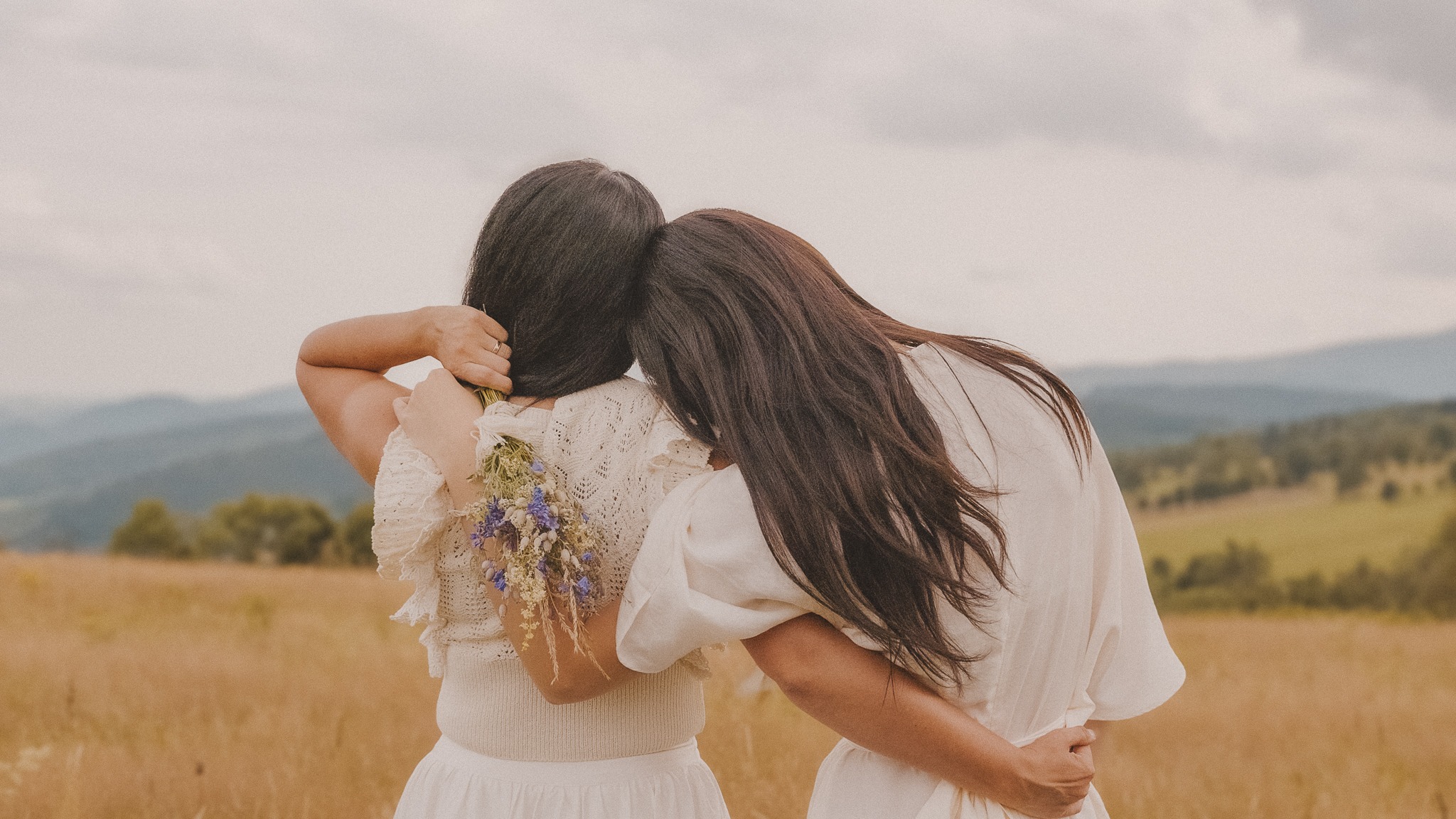 Dwie kobiety w jasnych sukienkach obejmują się na tle łąki i gór, jedna trzyma bukiet polnych kwiatów.