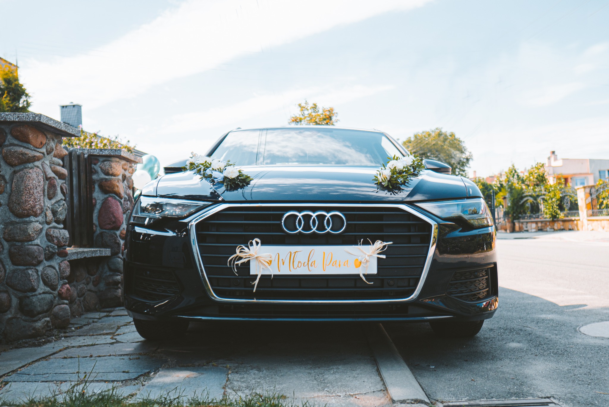 Czarny samochód Audi udekorowany kwiatami i tablicą 'Młoda Para' zaparkowany na zewnątrz, widok z przodu.