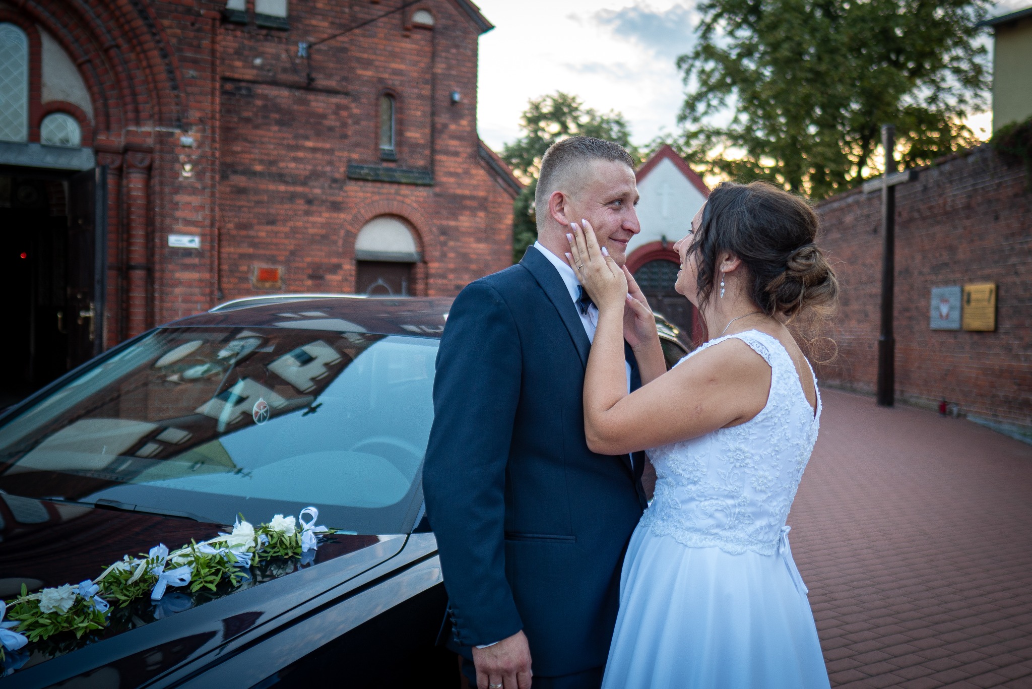Para młoda w eleganckich strojach stoi przed udekorowanym kwiatami samochodem na tle ceglanego budynku.