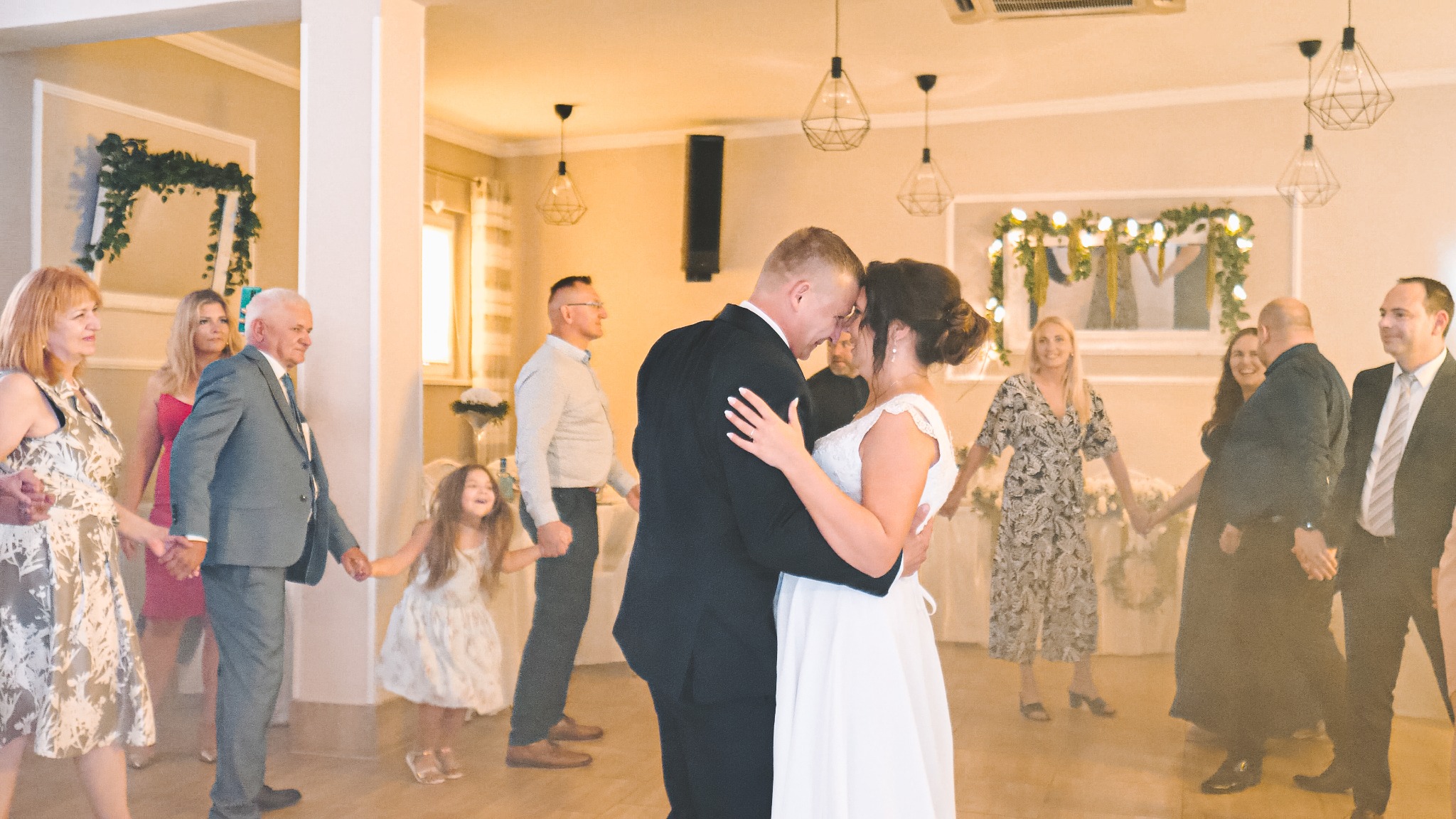 Para młoda podczas pierwszego tańca na sali weselnej, otoczona gośćmi trzymającymi się za ręce, dekoracje roślinne i geometryczne lampy wiszące.