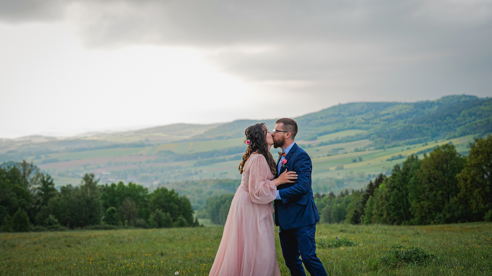 Pocałunek nowożeńców na łące z widokiem na zalesione wzgórza w pochmurny dzień, panna młoda w pudrowej sukni z kwiatami we włosach, pan młody w granatowym garniturze.