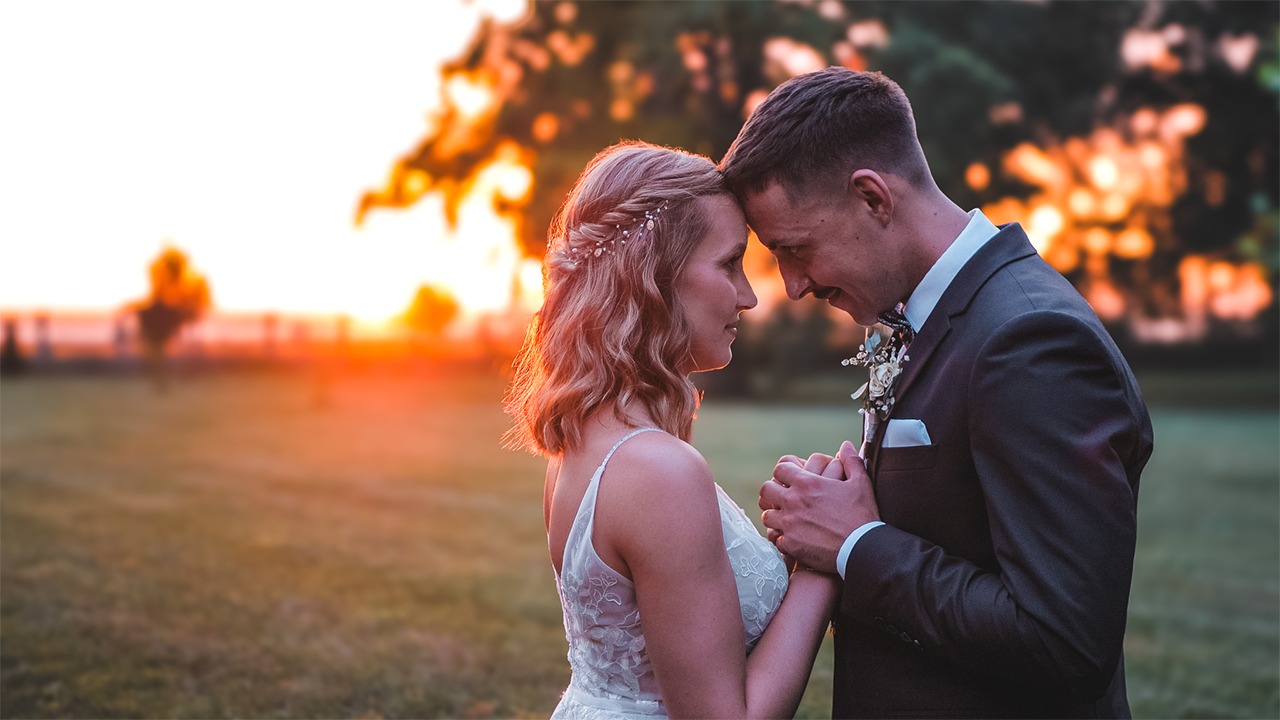 Para młoda, panna młoda w koronkowej sukni i pan młody w ciemnym garniturze, stoją blisko siebie czołami w zachodzącym słońcu na tle zieleni.