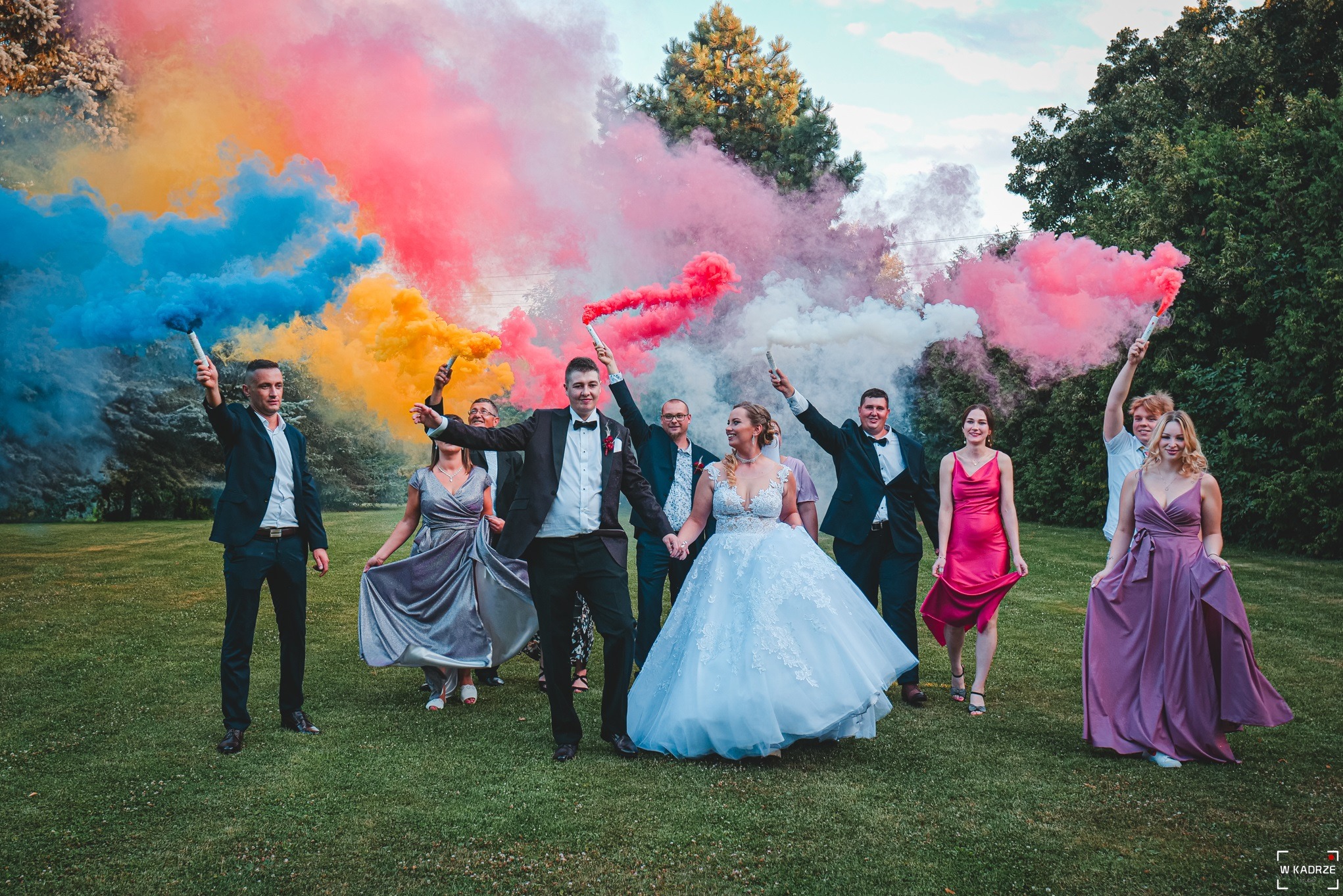 Para młoda otoczona gośćmi weselnymi z kolorowymi racami dymnymi na tle zielonej trawy i drzew.