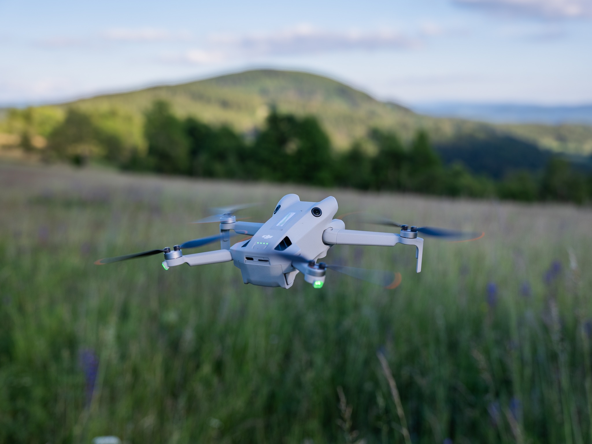 Szary dron w locie na tle zielonego wzgórza i łąki z fioletowymi kwiatami.