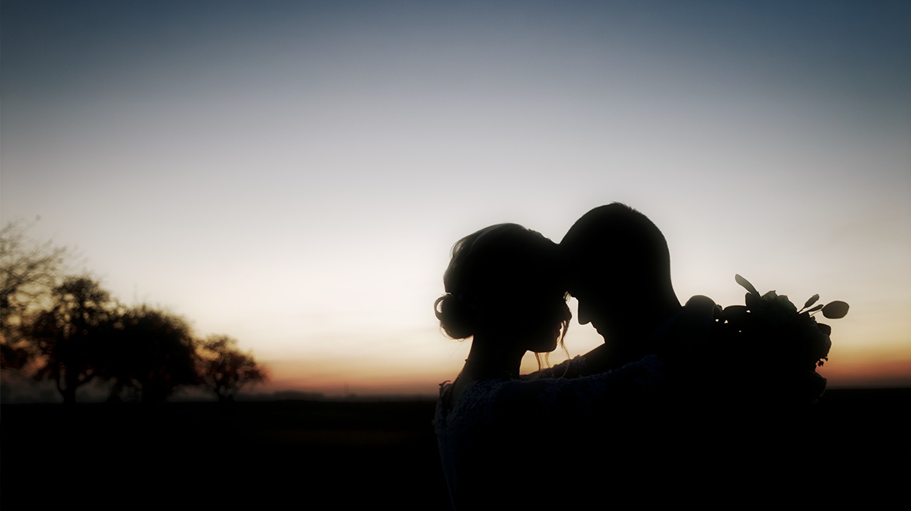 Sylwetki pary młodej obejmującej się na tle zachodzącego słońca z bukietem ślubnym, widoczne drzewa w oddali.