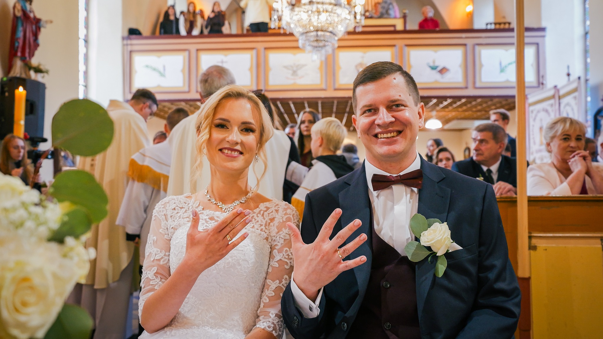 Uśmiechnięta para młoda w kościele prezentuje obrączki, na drugim planie goście i księża.