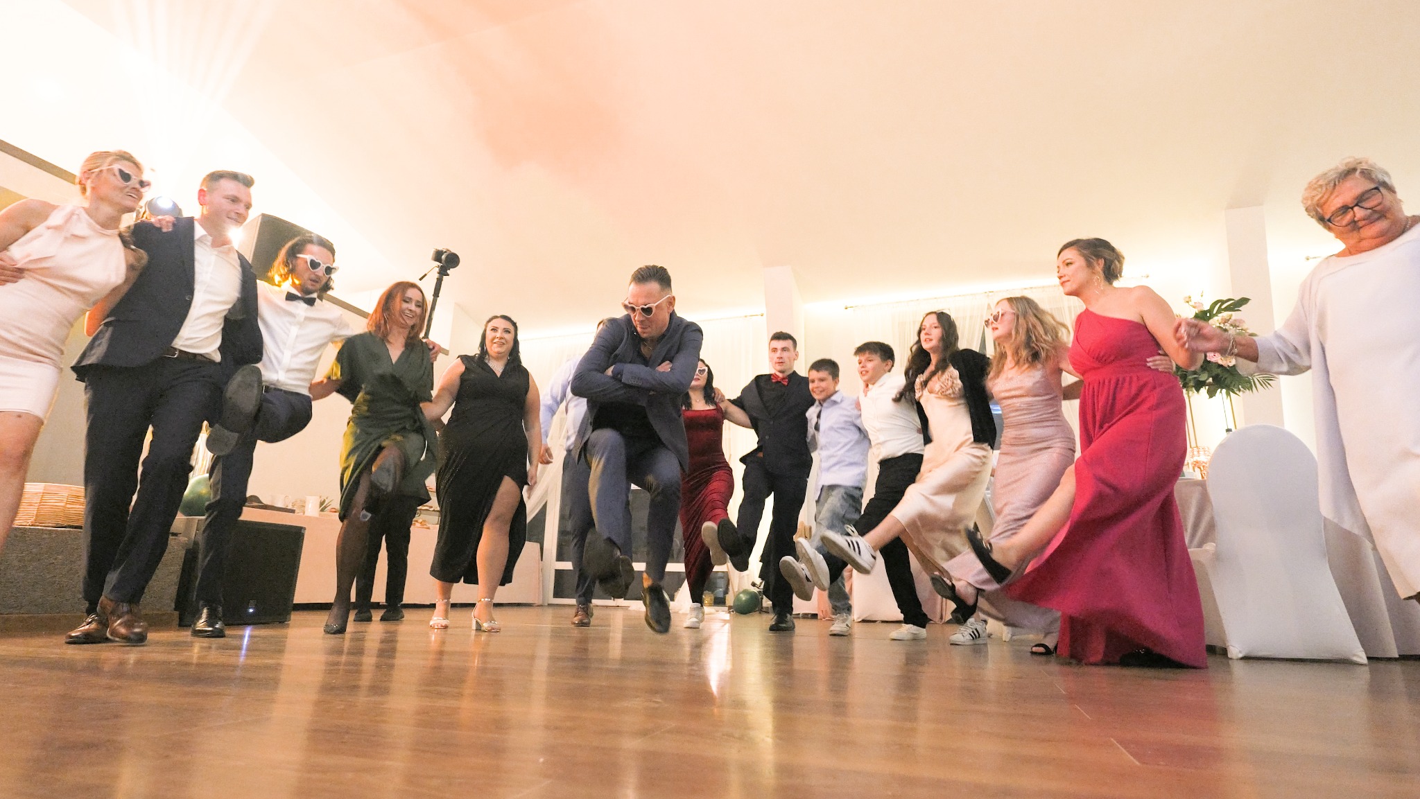 Grupa osób w eleganckich strojach, tańcząca w rzędzie na parkiecie podczas uroczystości, z widocznym oświetleniem sufitowym i elementami dekoracji.