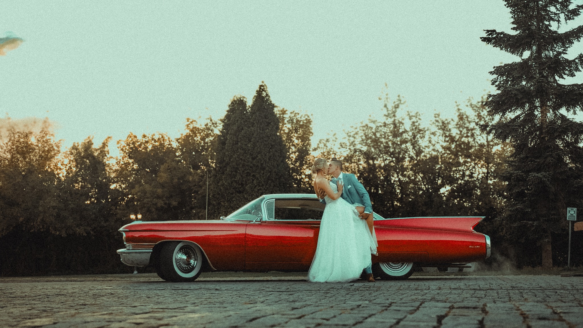 Pocałunek nowożeńców przy czerwonym, klasycznym Cadillacu Eldorado z białym dachem na tle drzew i nieba o zachodzie słońca.