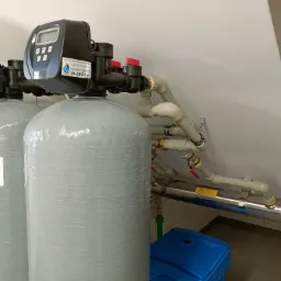Dwa szare zbiorniki z elektronicznymi panelami sterowania i rurami, prawdopodobnie system filtracji wody, zainstalowane w pomieszczeniu z białymi ścianami i szarą podłogą. Na zbiorniku widoczna...