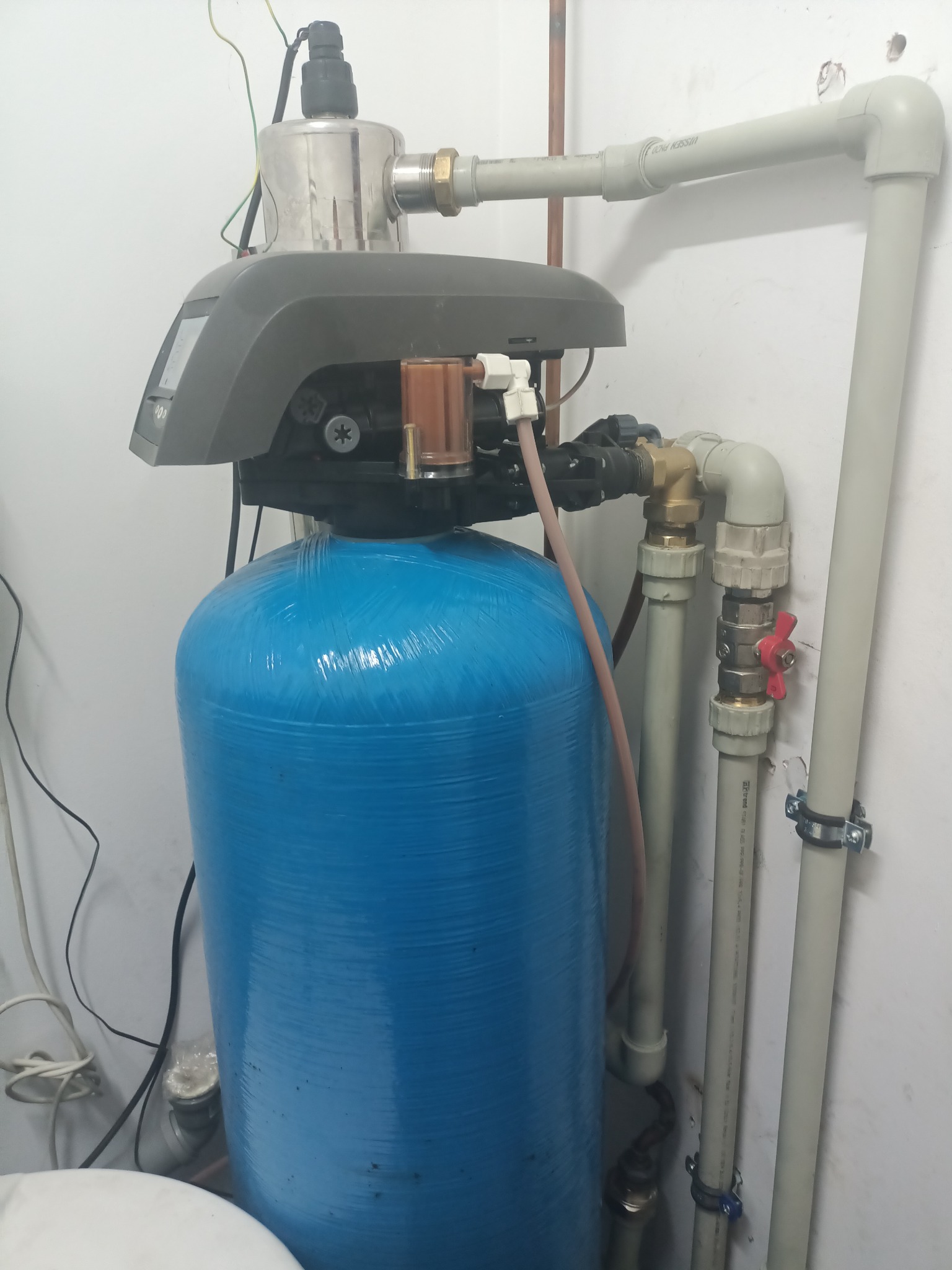 Niebieski zbiornik stacji uzdatniania wody z szarą głowicą sterującą i orurowaniem PVC w pomieszczeniu technicznym.