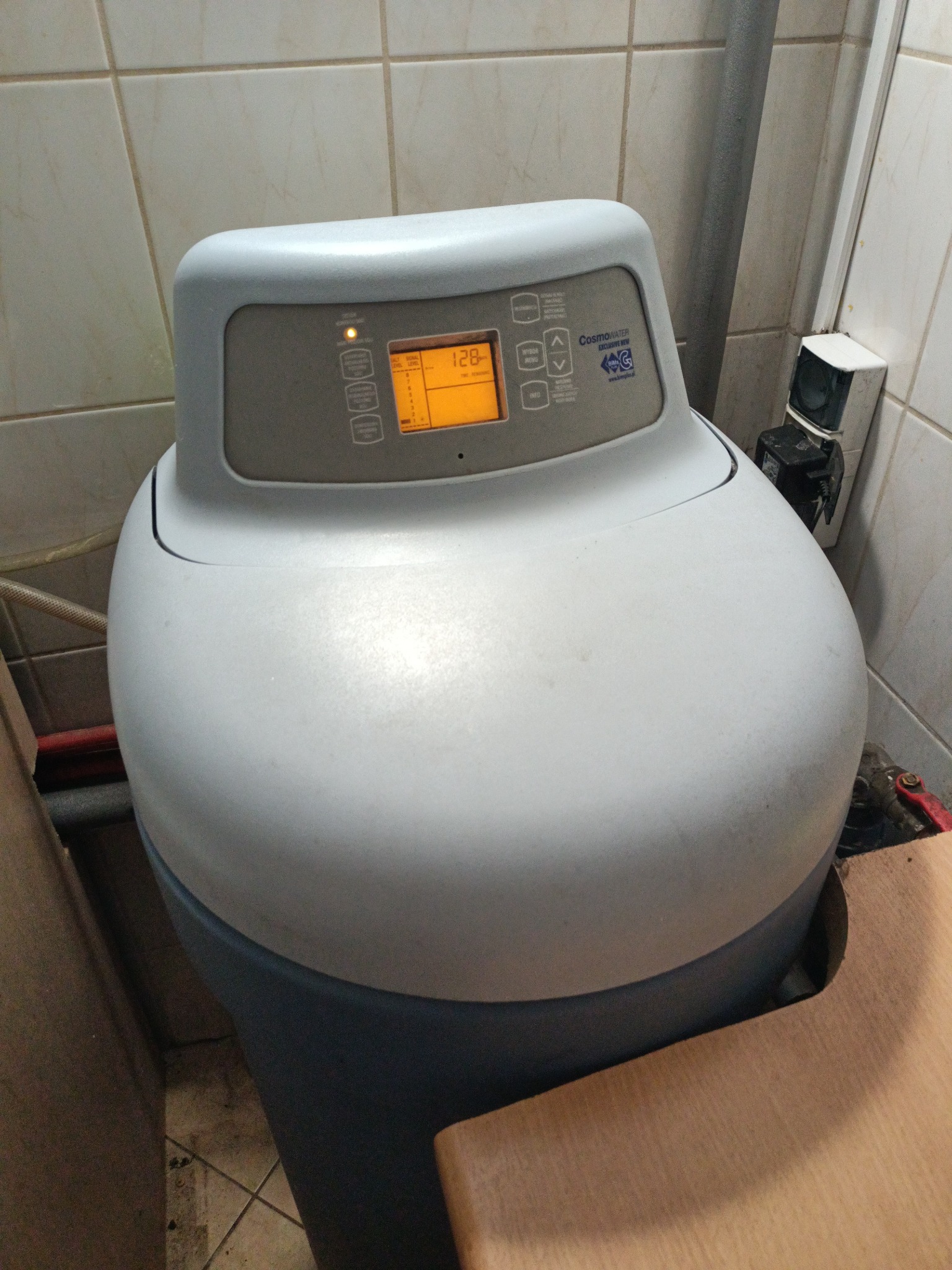 Szaro-niebieskie urządzenie do zmiękczania wody w łazience z beżowymi płytkami, widoczny wyświetlacz z pomarańczowymi cyframi i logo producenta.