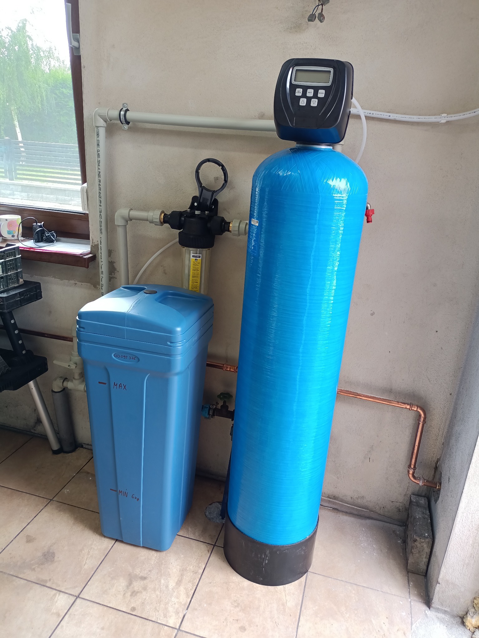 Niebieski system filtracji wody z wyświetlaczem cyfrowym, obok zbiornik na sól, instalacja z rur miedzianych, widoczne okno i fragment pomieszczenia.
