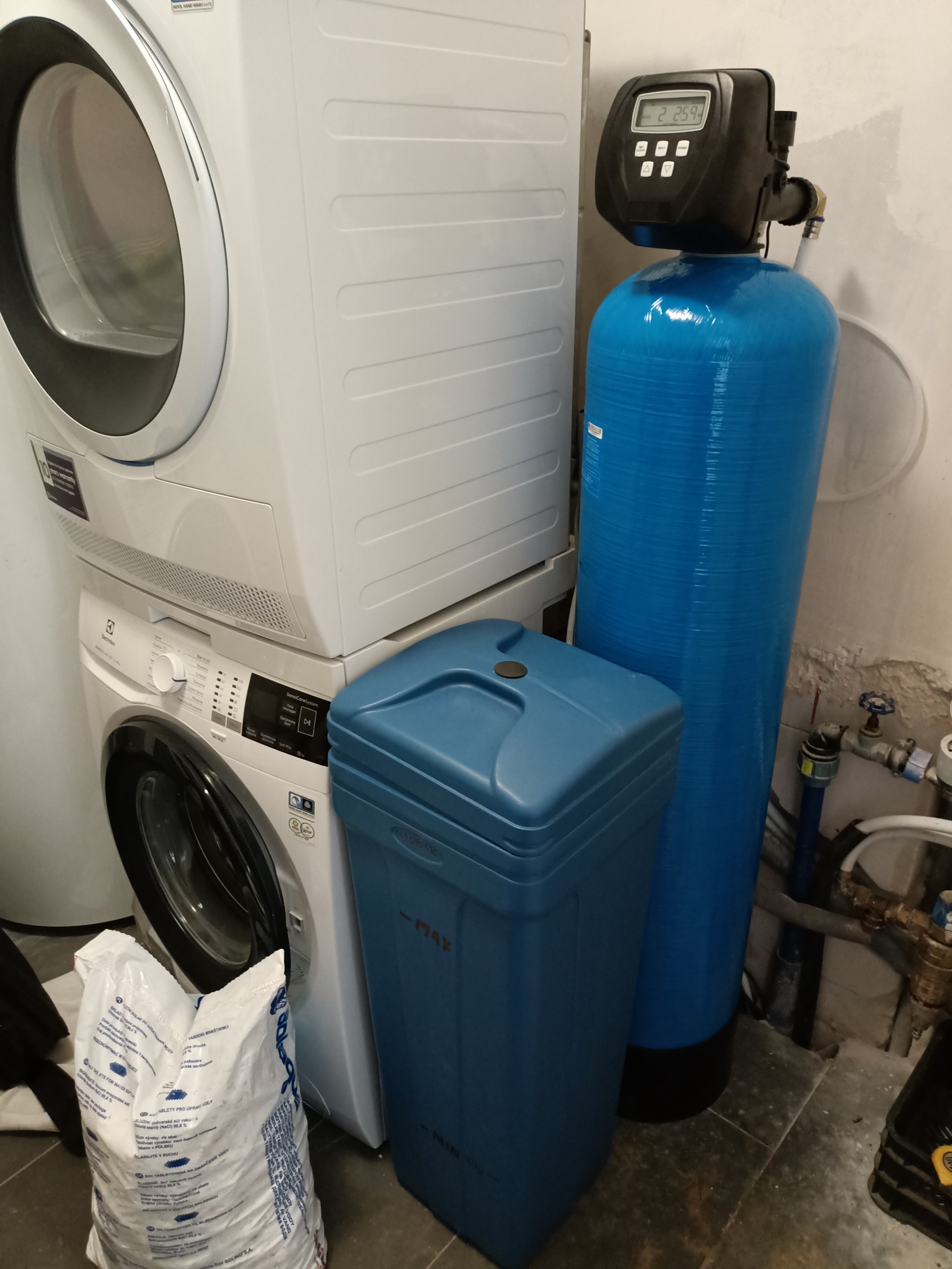 Domowa stacja uzdatniania wody z niebieskim zbiornikiem i elektronicznym sterownikiem, obok pralka i suszarka, worek z solą tabletkowaną do zmiękczania wody.