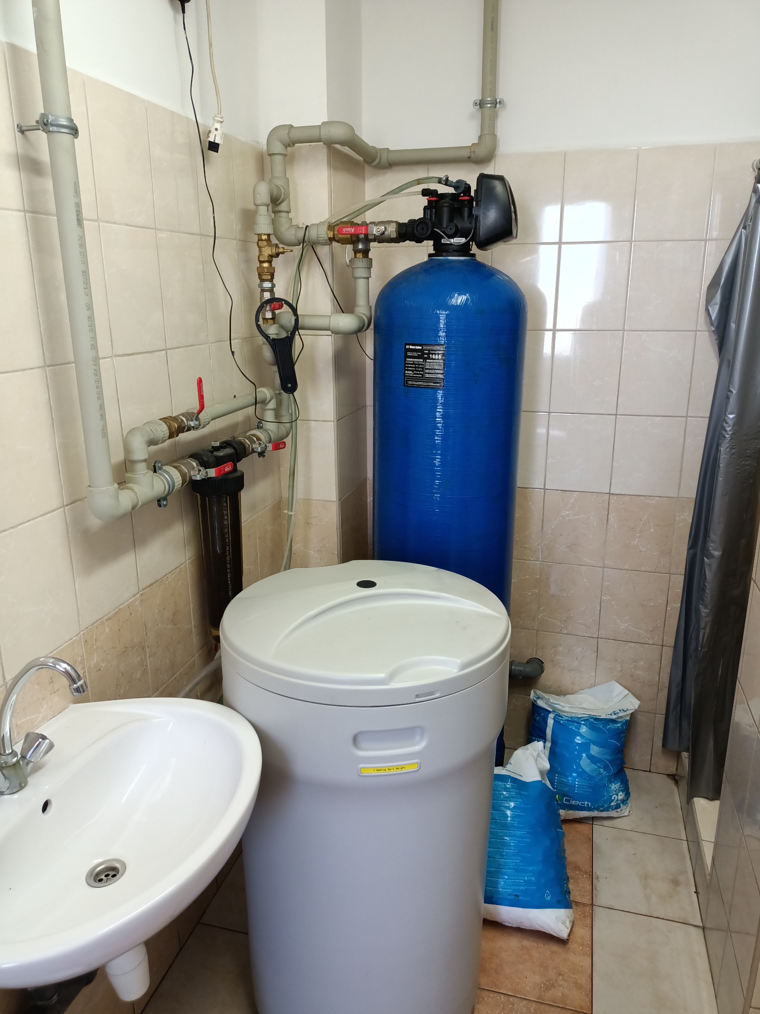 Domowa instalacja uzdatniania wody z niebieskim zbiornikiem, białym zmiękczaczem i workami soli regeneracyjnej w kafelkowanej łazience, widoczna umywalka i elementy instalacji wodnej.