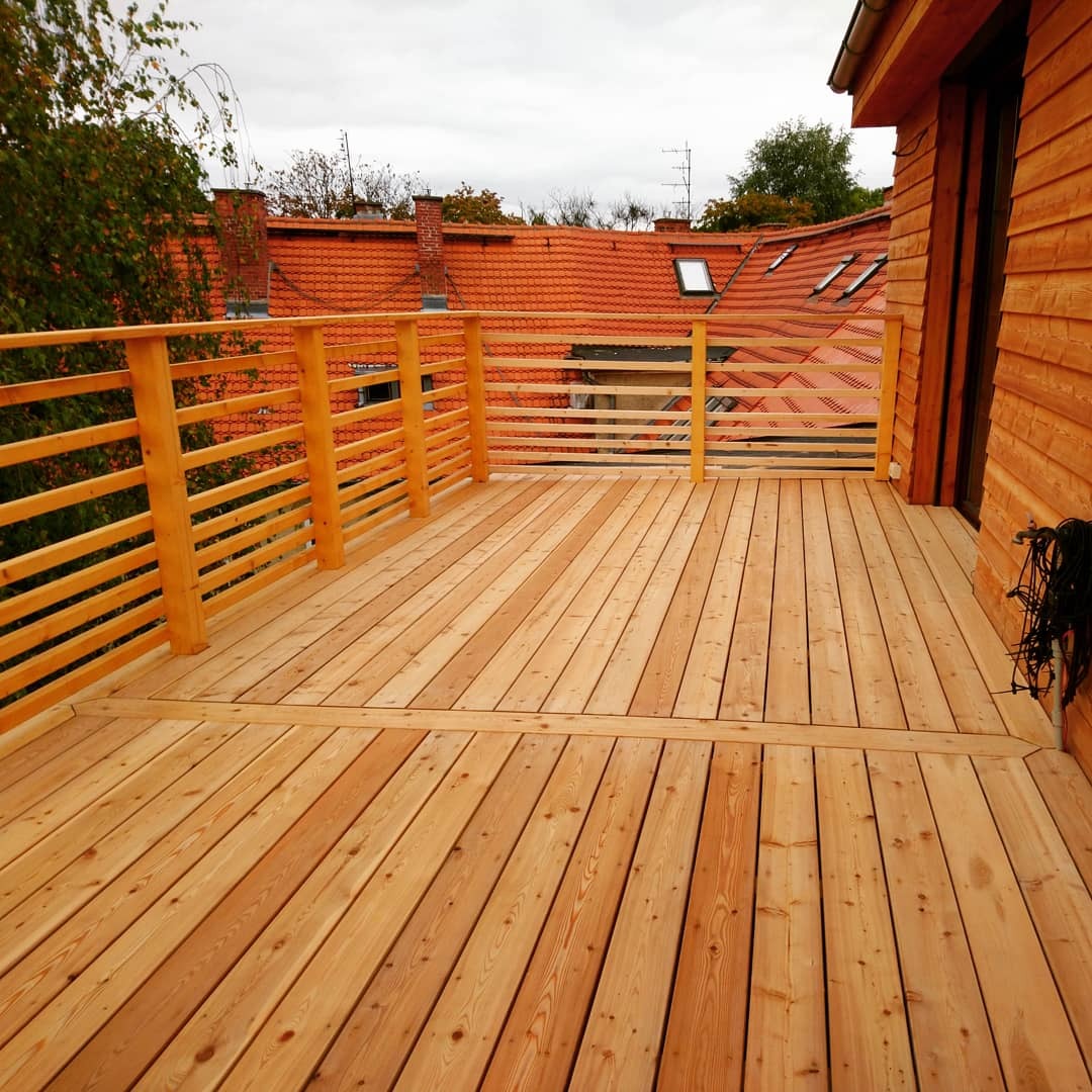 Drewniany taras z balustradą, widok na dachy budynków z czerwonej dachówki i fragment drzewa. Jasne drewno, proste linie balustrady, pochmurne niebo w tle.