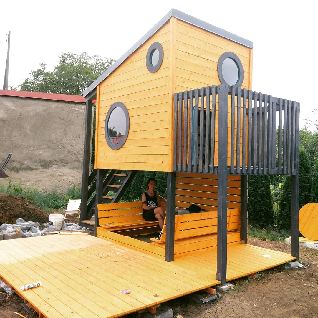 Drewniany domek dla dzieci z okrągłymi oknami, balkonem i tarasem, pomalowany na żółto, z szarymi elementami konstrukcyjnymi; kobieta siedzi w środku. Częściowo ukończony, widoczne ślady budowy...