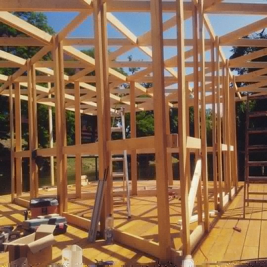 Konstrukcja szkieletowa drewnianego budynku w trakcie budowy, widoczne narzędzia i drabiny, otoczenie zielone.