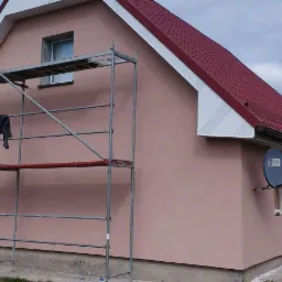 Dom jednorodzinny z nową, jednolitą elewacją w odcieniu pudrowego r&oacute;żu, z widocznym rusztowaniem i fragmentem czerwonego dachu. Na ścianie zamontowana antena satelitarna.