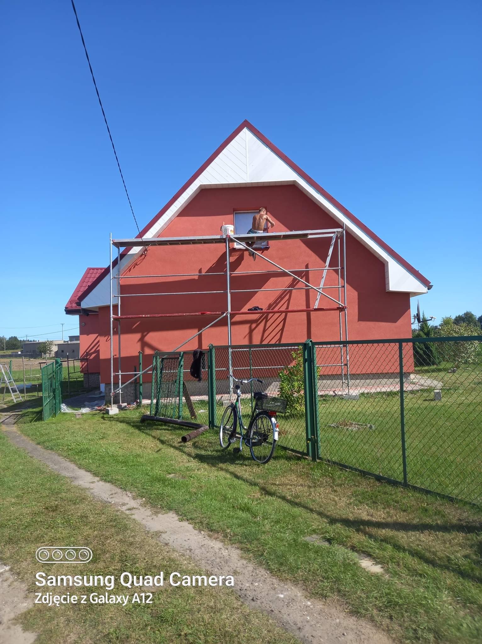 Mężczyzna bez koszulki na rusztowaniu, malujący górną część czerwonej elewacji domu z białymi elementami wykończeniowymi, widoczny zza zielonego ogrodzenia, obok którego stoi rower.