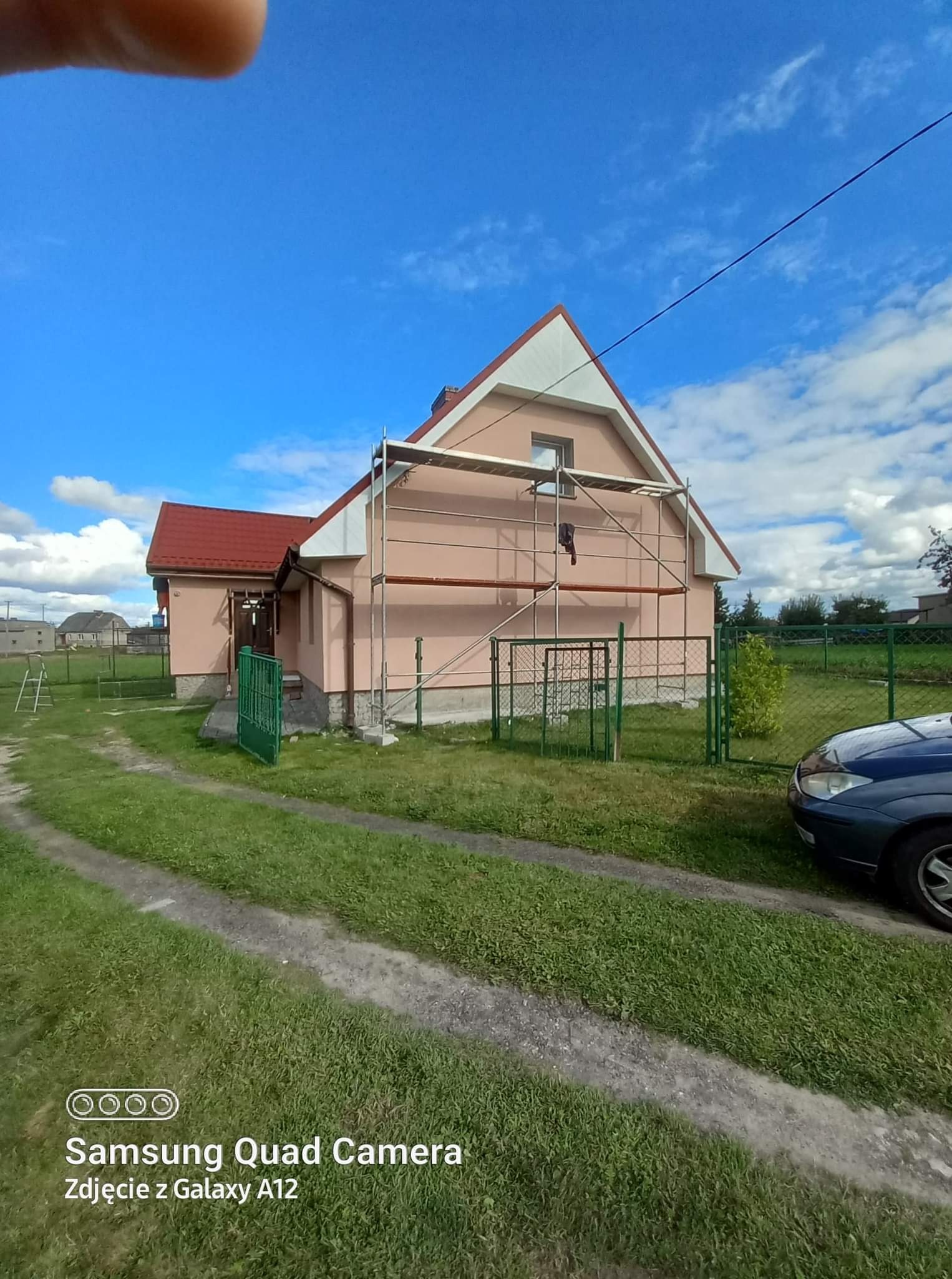 Różowy dom jednorodzinny w trakcie renowacji elewacji, widoczne rusztowanie, częściowo odnowiona ściana, zielony trawnik i błękitne niebo.