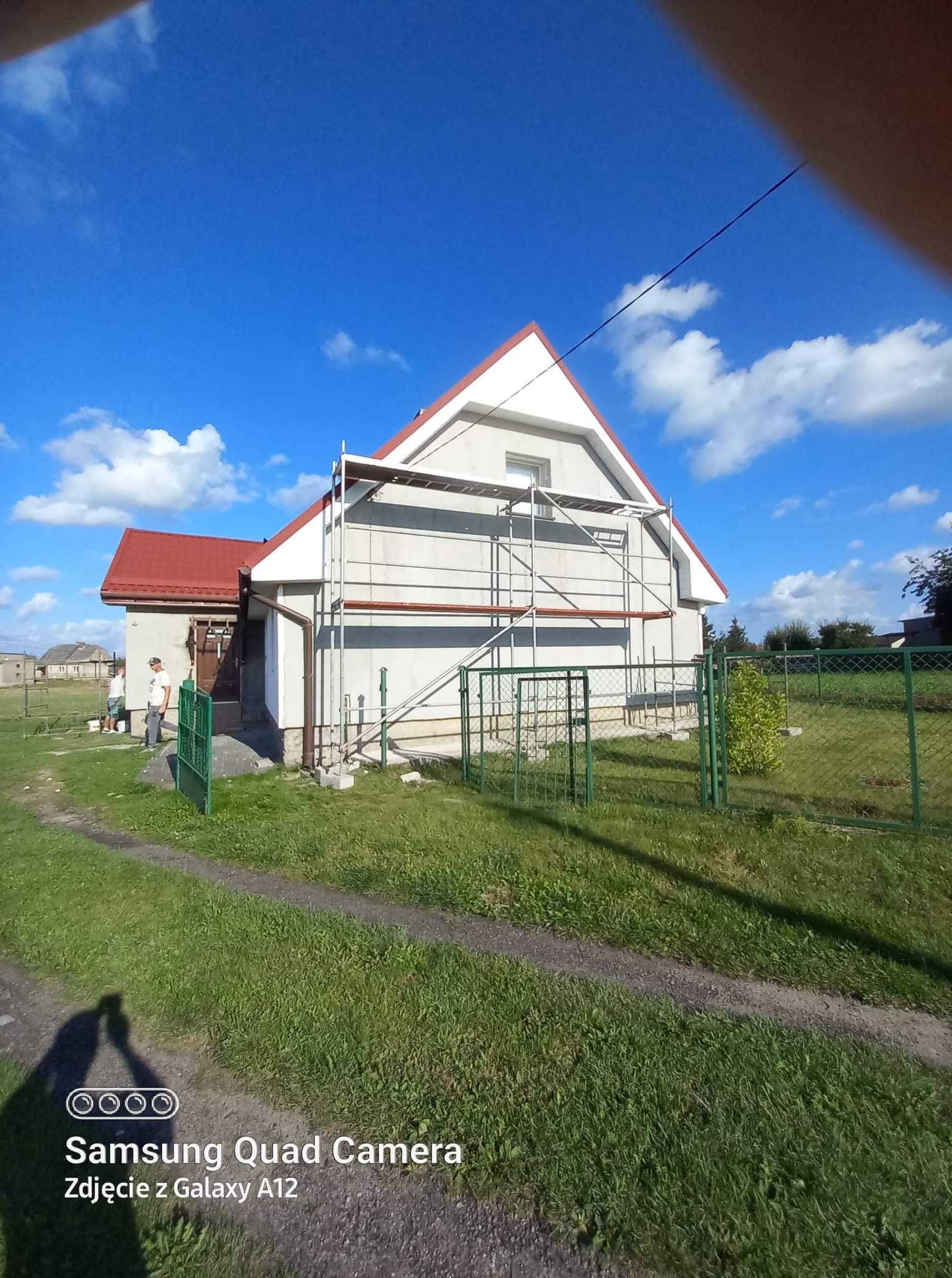 Dom jednorodzinny w trakcie prac elewacyjnych, widoczne rusztowanie, częściowo pomalowana fasada, trawnik, zielone ogrodzenie i błękitne niebo z chmurami.