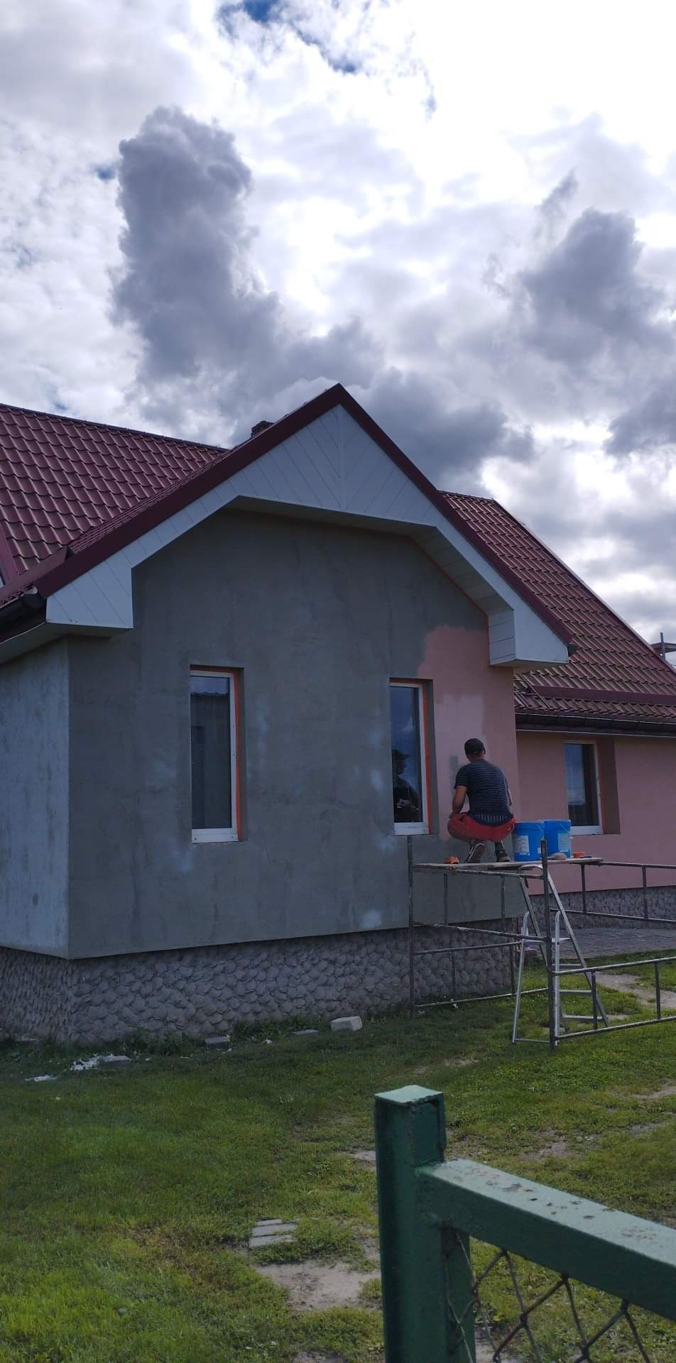 Mężczyzna na rusztowaniu maluje elewację domu na kolor różowy, widoczne dwa okna z pomarańczowymi ramami, część ściany w szarym tynku, dach z czerwonej dachówki, pochmurne niebo.