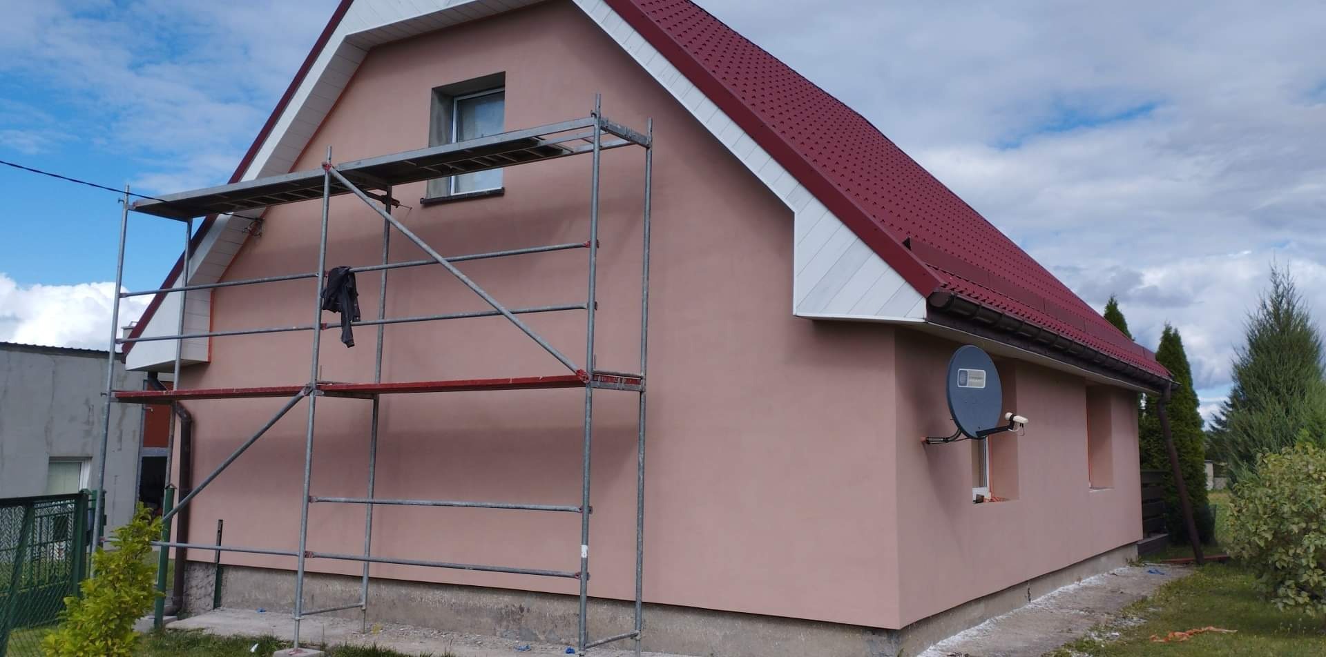 Dom jednorodzinny z nową, jednolitą elewacją w odcieniu pudrowego różu, z widocznym rusztowaniem i fragmentem czerwonego dachu. Na ścianie zamontowana antena satelitarna.