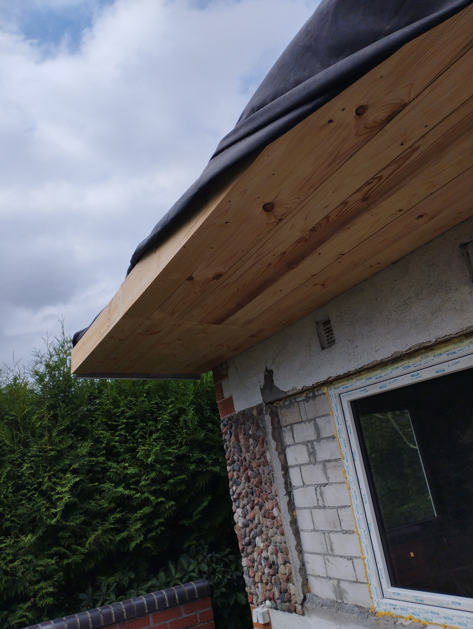 Drewniana konstrukcja okapu dachu zabezpieczona czarną folią, widoczna na tle pochmurnego nieba i fragmentu elewacji z oknem oraz zielenią.