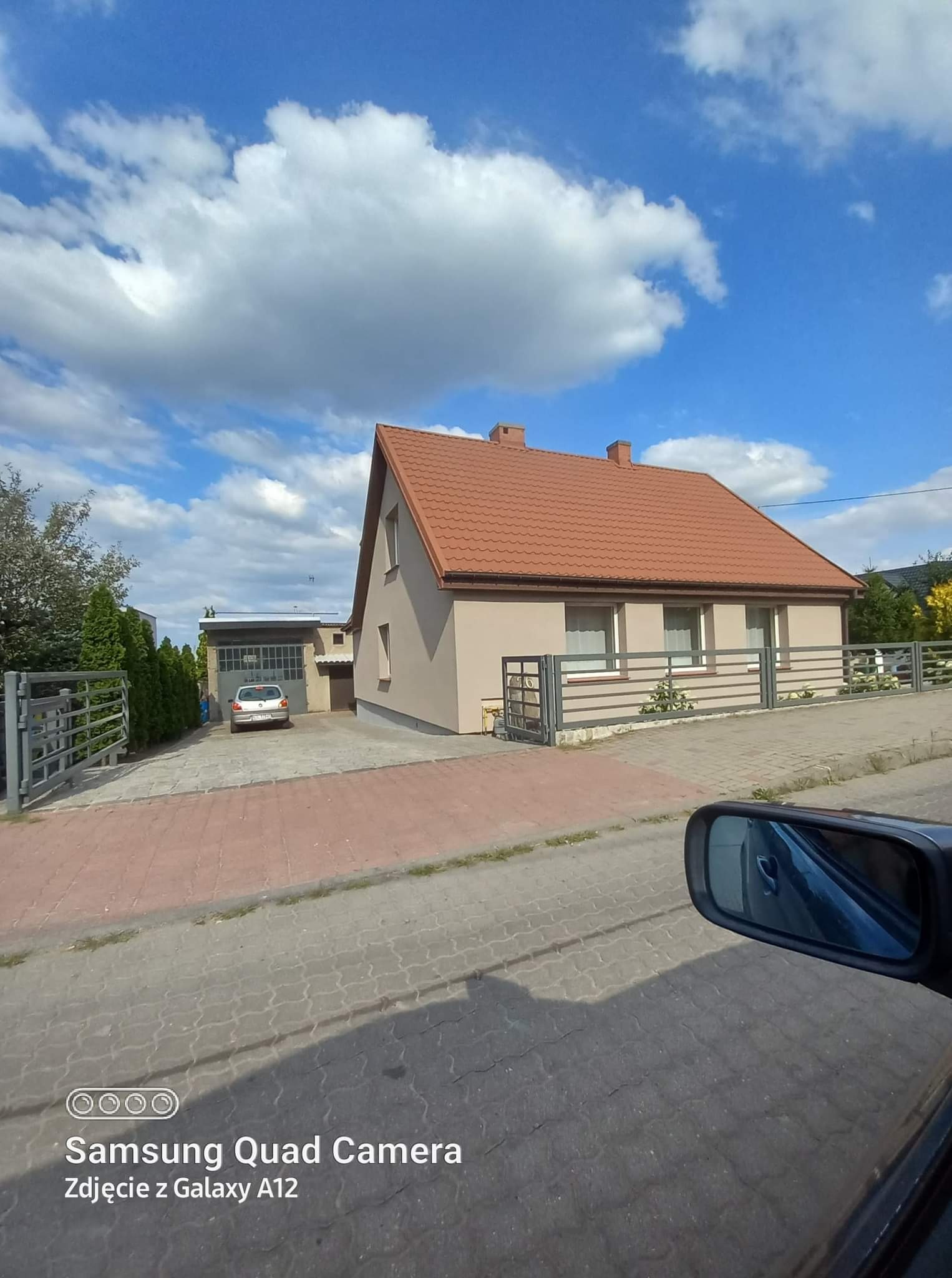 Jasny, jednopiętrowy dom z brązowym dachem, widoczny z perspektywy ulicy, z fragmentem lusterka samochodowego w prawym dolnym rogu i garażem w tle.