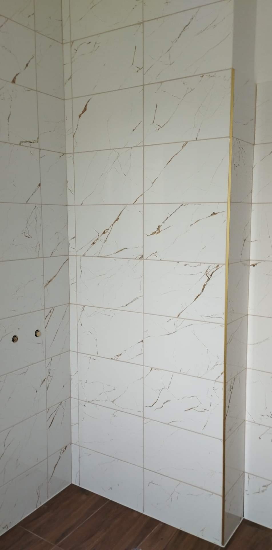 Wykończona ściana łazienki z białymi płytkami imitującymi marmur z brązowymi żyłkami, widoczne dwa otwory na armaturę i brązowa podłoga imitująca drewno.