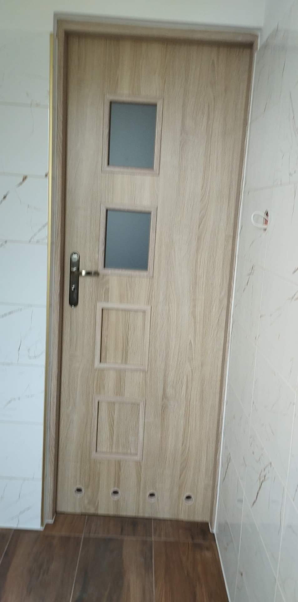 Nowe drzwi wewnętrzne z czterema kwadratowymi panelami, dwoma szklanymi i dwoma pełnymi, zamontowane w łazience z wykończoną glazurą imitującą marmur i brązową podłogą z płytek.