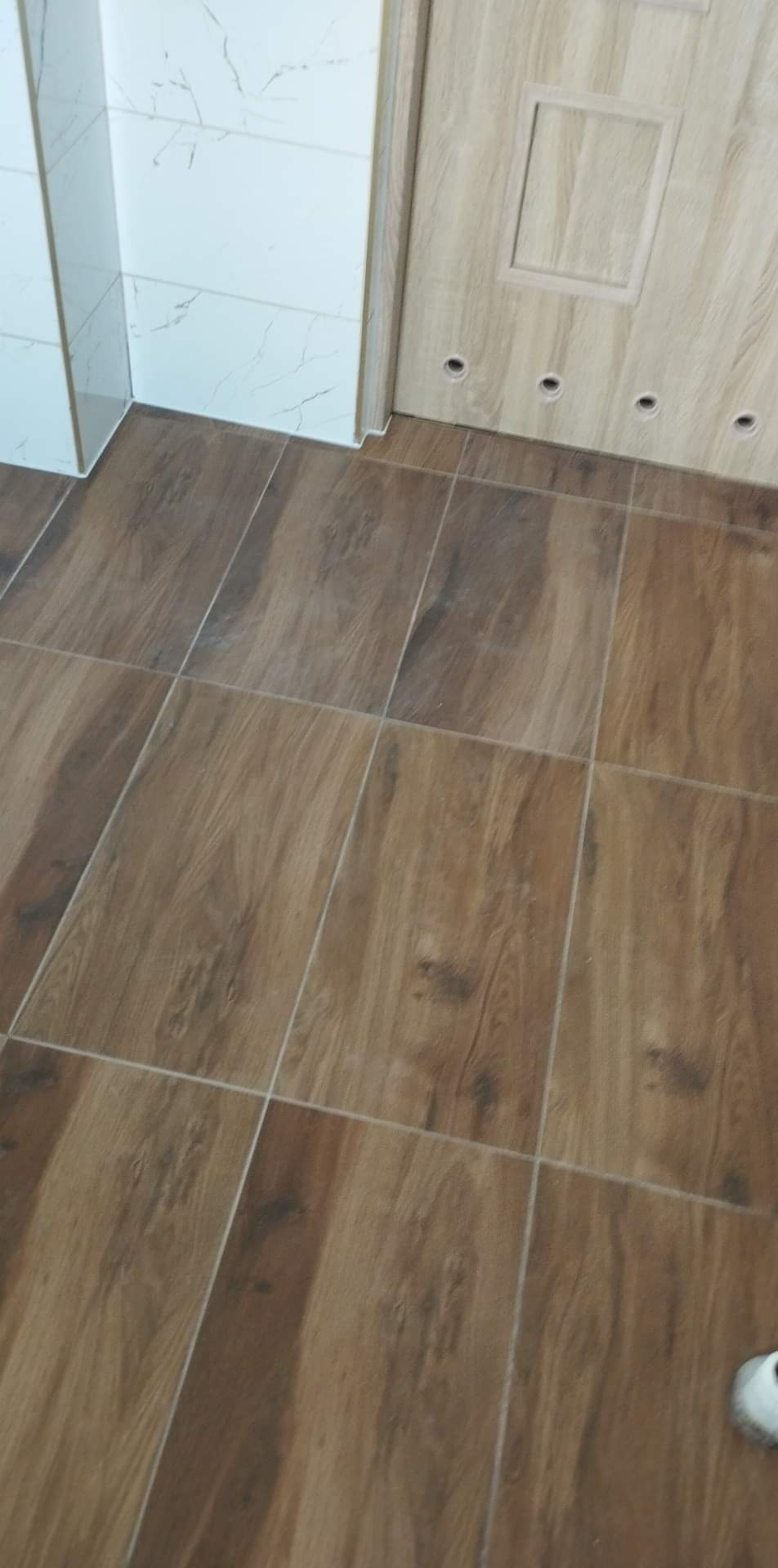 Brązowe płytki podłogowe imitujące drewno, ułożone w łazience, z widocznym fragmentem białych płytek ściennych z szarym wzorem marmuru oraz dolną częścią drewnianej szafki z otworami.