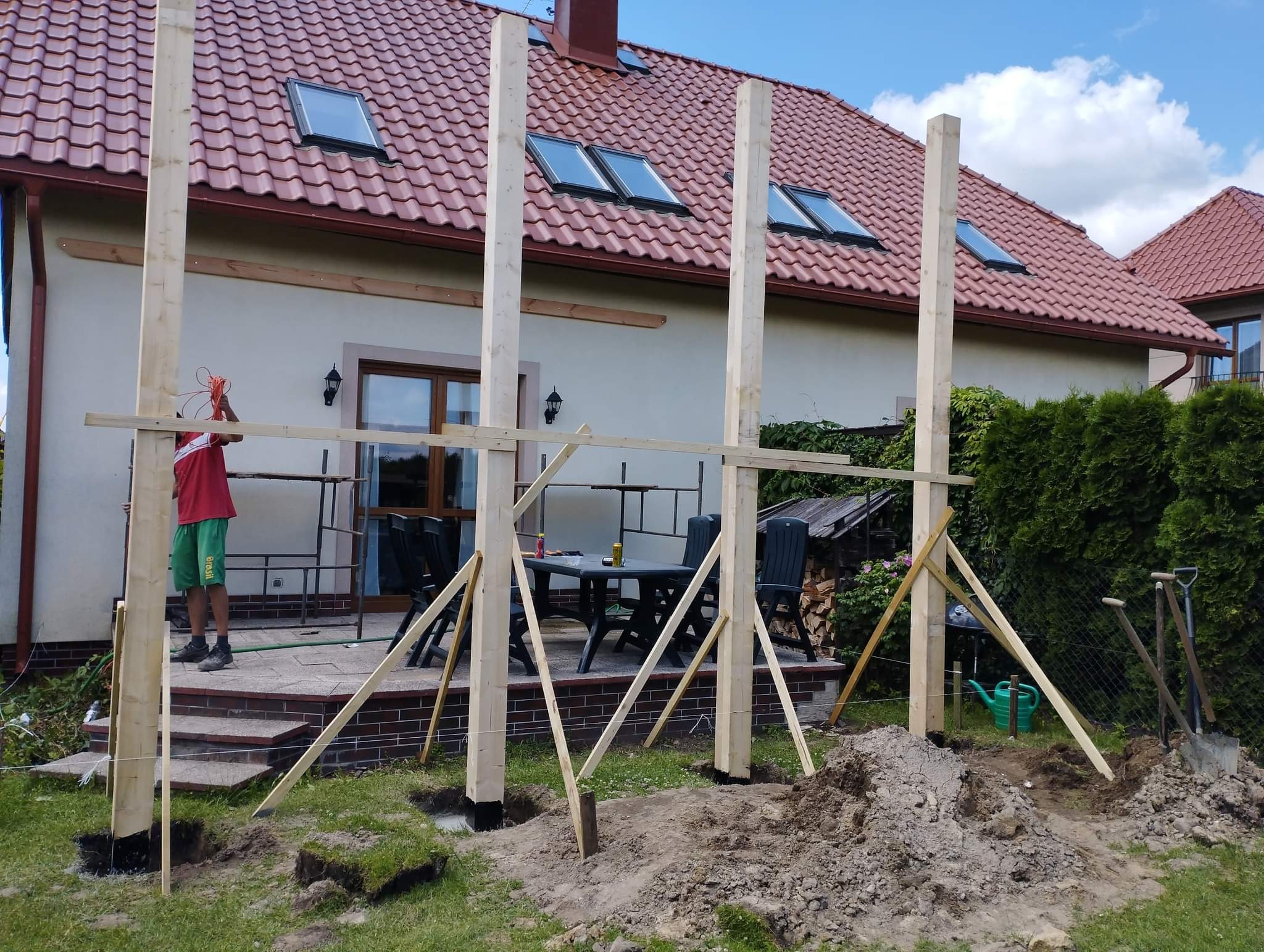 Montaż drewnianej konstrukcji z belek na tarasie domu jednorodzinnego, widoczne słupy, podpory, liny pomiarowe oraz pracownik z pomarańczowym przewodem.