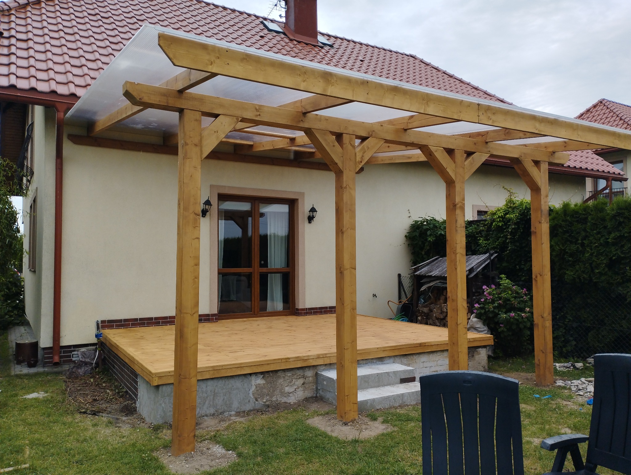 Drewniana pergola z przezroczystym zadaszeniem nad tarasem z desek, przylegająca do domu z beżową elewacją i czerwoną dachówką.