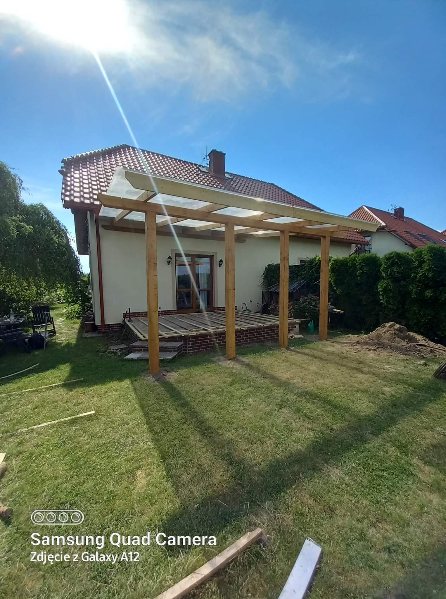 Drewniana pergola z przezroczystym zadaszeniem w trakcie budowy, widoczne słupy i konstrukcja nośna, częściowo ułożona podłoga tarasu na podwyższeniu z cegły, dom jednorodzinny w tle, słoneczny dzień.