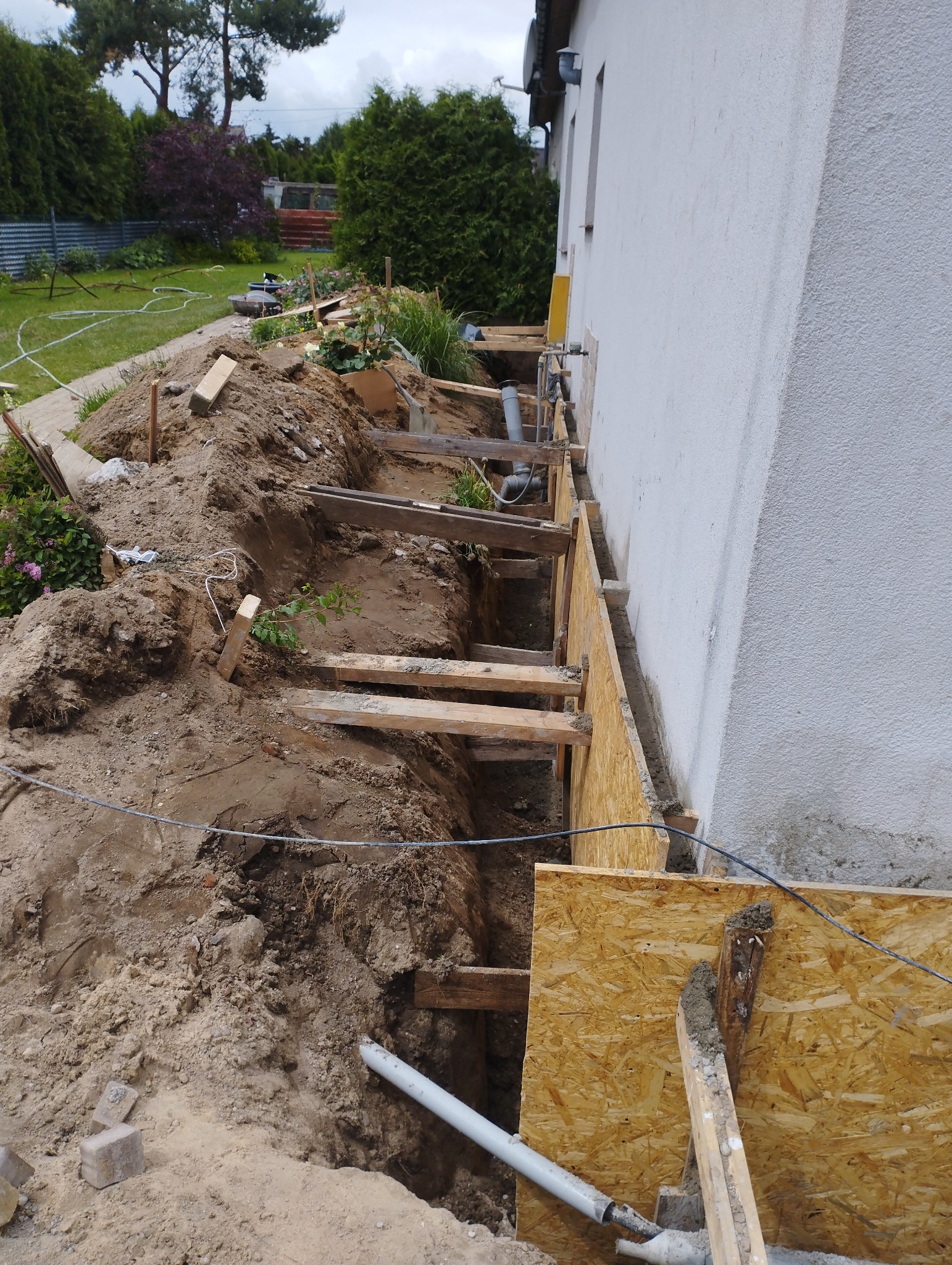 Wykop fundamentów przy ścianie budynku, wzmocniony drewnianą konstrukcją i płytami OSB, widoczne rury kanalizacyjne, ziemia i elementy ogrodu w tle.