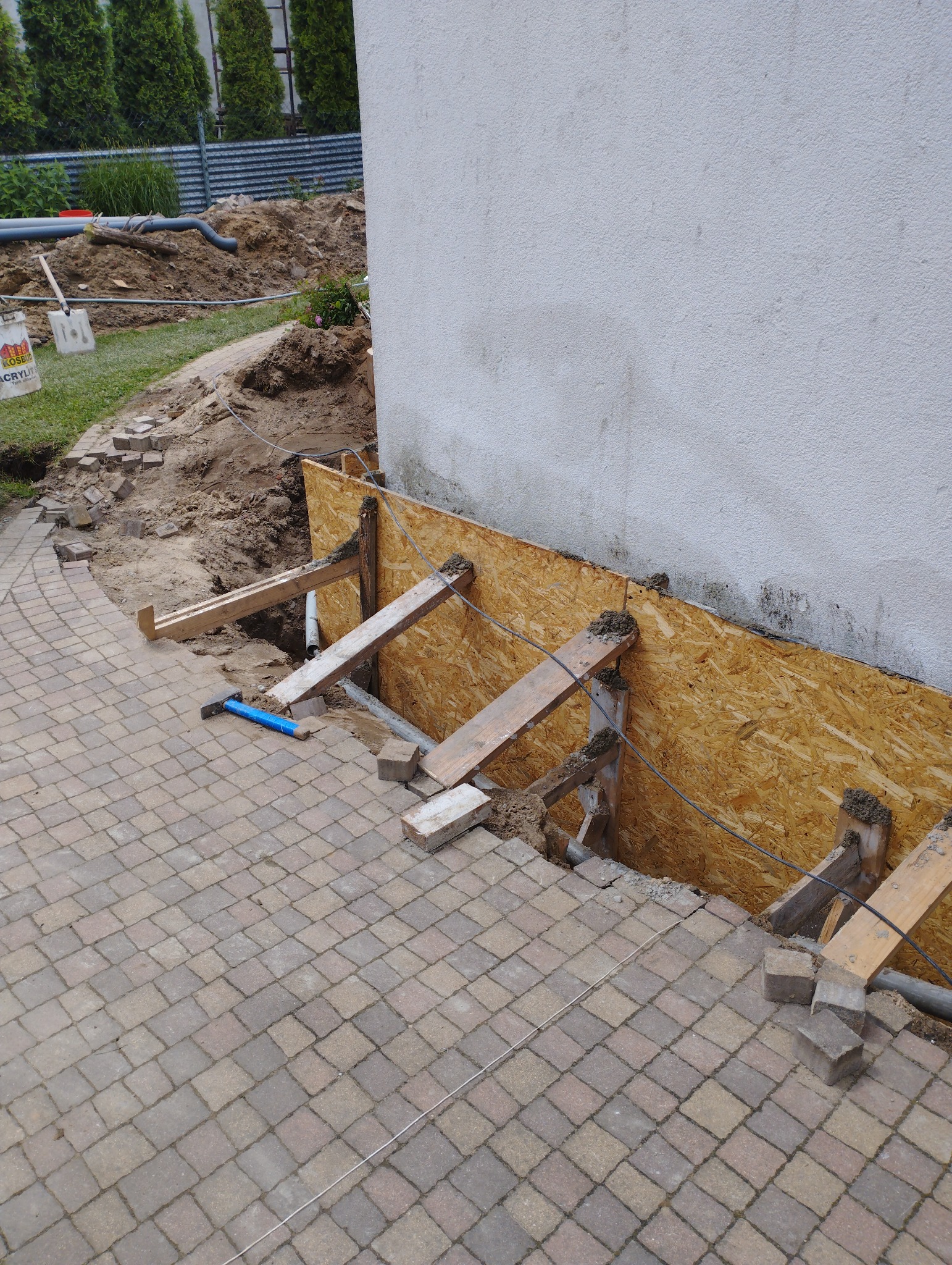 Przygotowanie do izolacji fundamentów budynku: deskowanie z płyty OSB wzmocnione drewnianymi belkami, widoczny wykop wzdłuż ściany, kostka brukowa i narzędzia w tle.