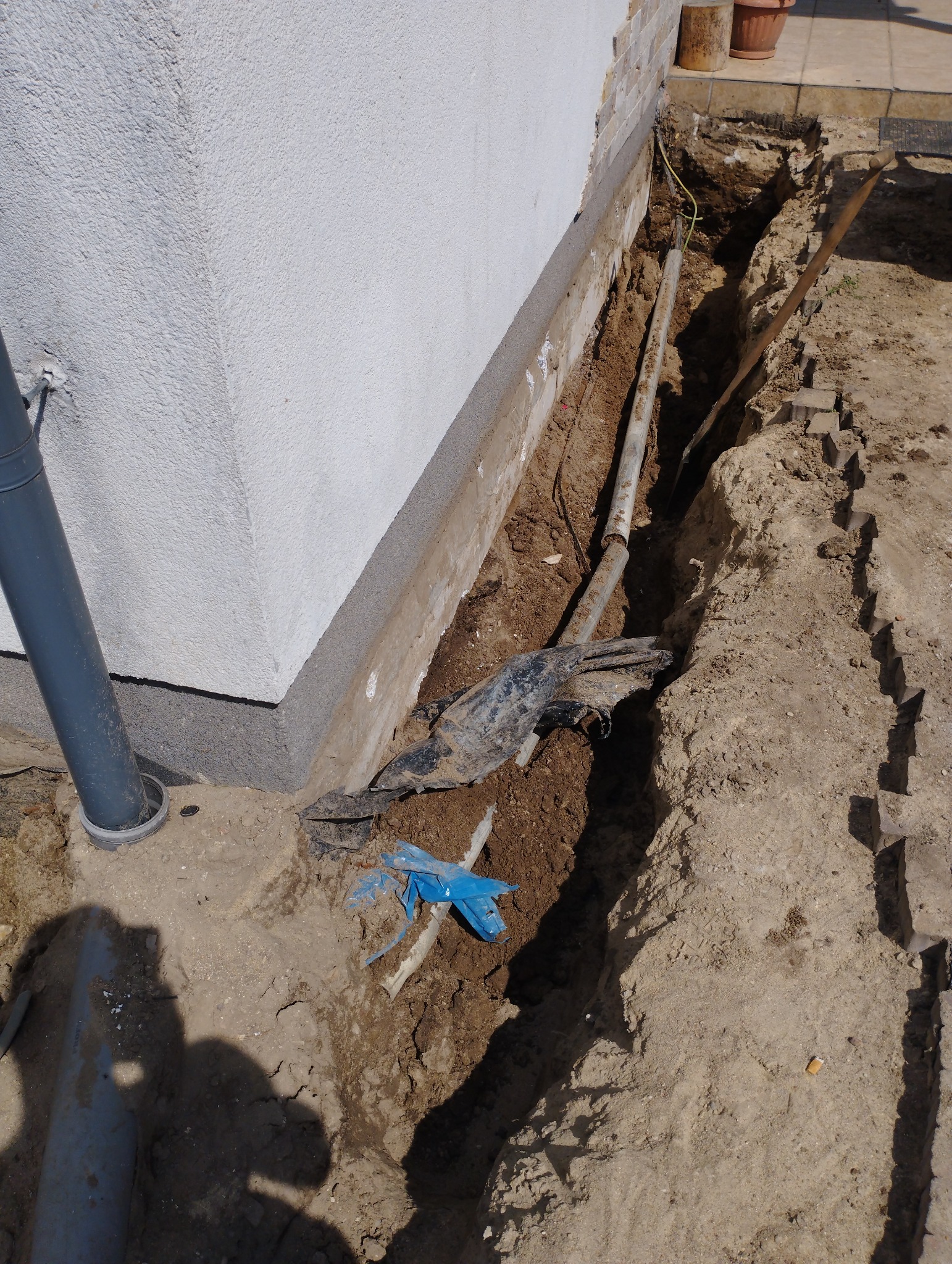 Wykop wzdłuż ściany budynku z widocznymi rurami, kablem elektrycznym i fragmentami folii, przygotowany pod prace elewacyjne.