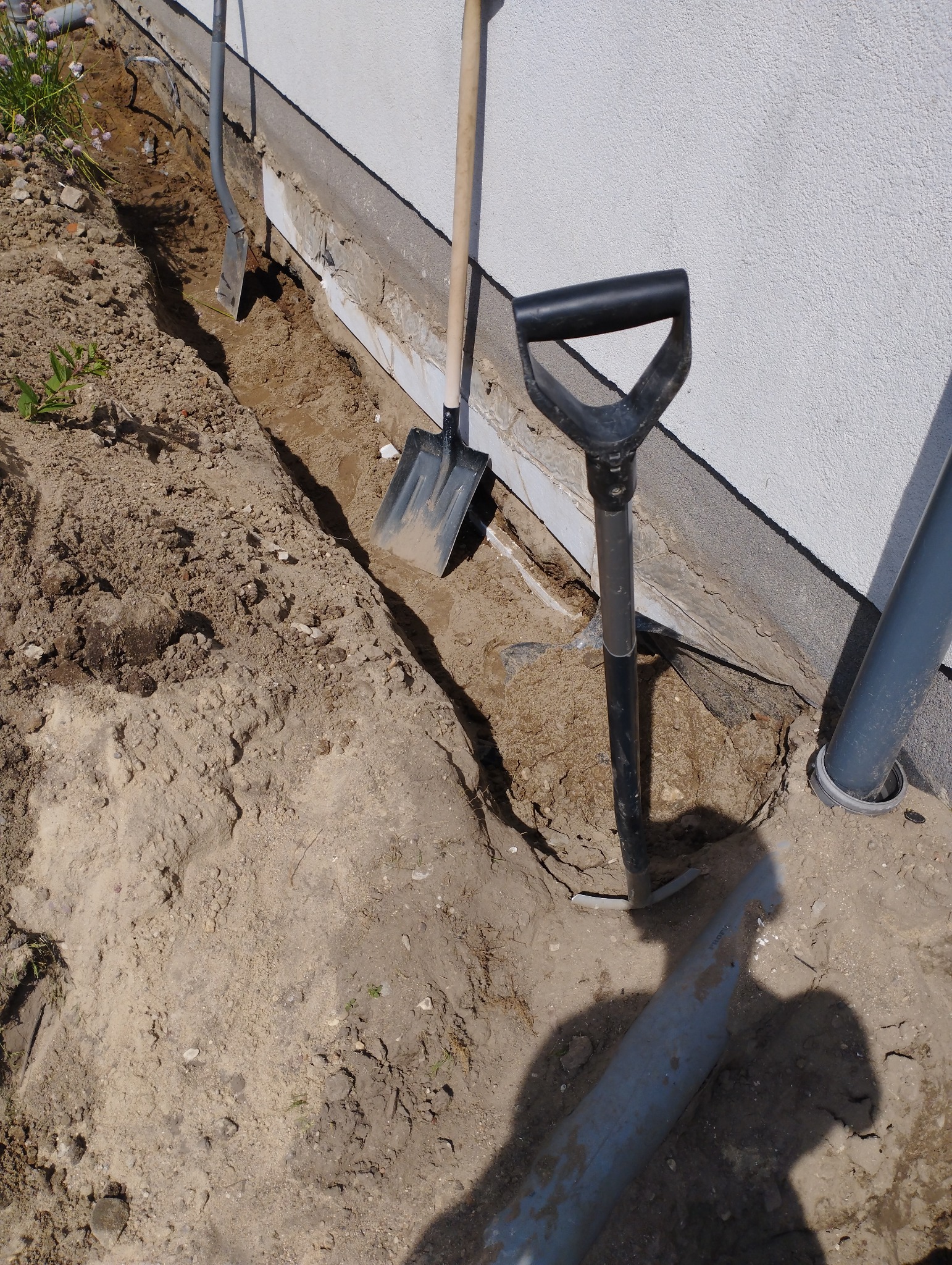 Wykop wzdłuż fundamentu budynku z widocznymi trzema szpadlami i fragmentem rury kanalizacyjnej, ukazujący przygotowania do prac elewacyjnych lub izolacyjnych.