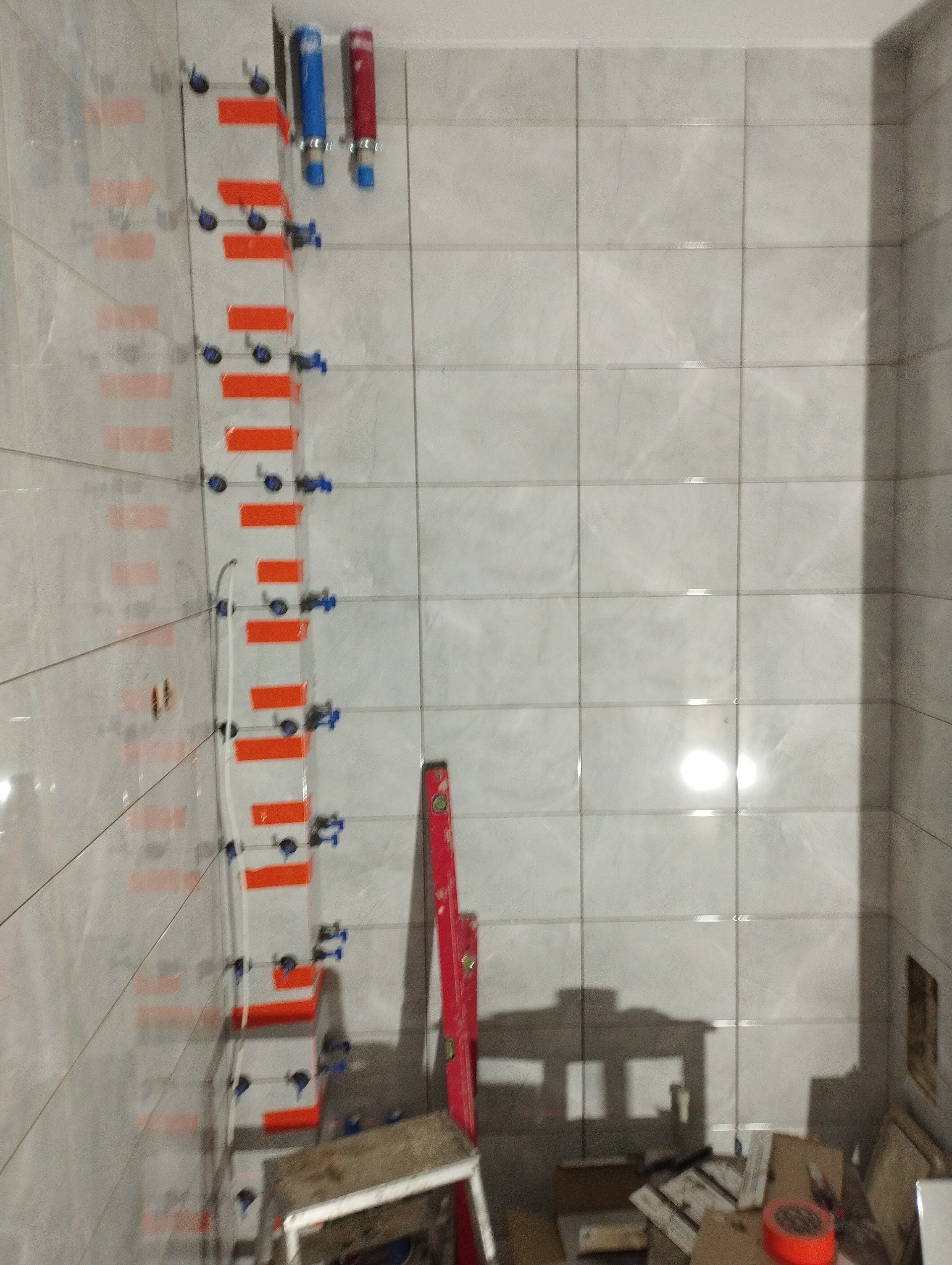Ściana łazienki w trakcie układania płytek ceramicznych z użyciem poziomicy i klipsów dystansowych, widoczne rury instalacji wodnej.