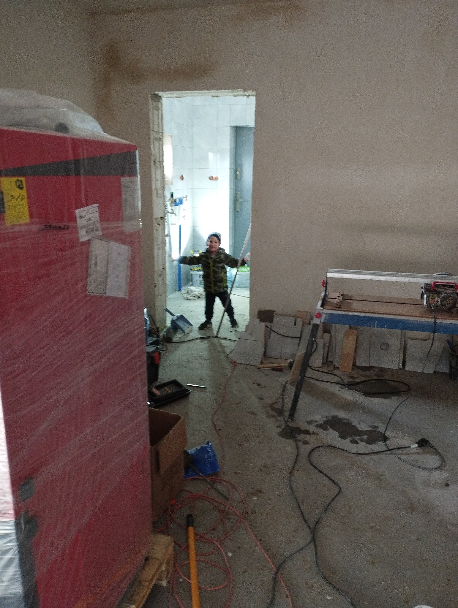 Wnętrze remontowanej łazienki z widoczną maszyną do cięcia płytek, materiałami budowlanymi i chłopcem w otwartych drzwiach do wykafelkowanej łazienki.