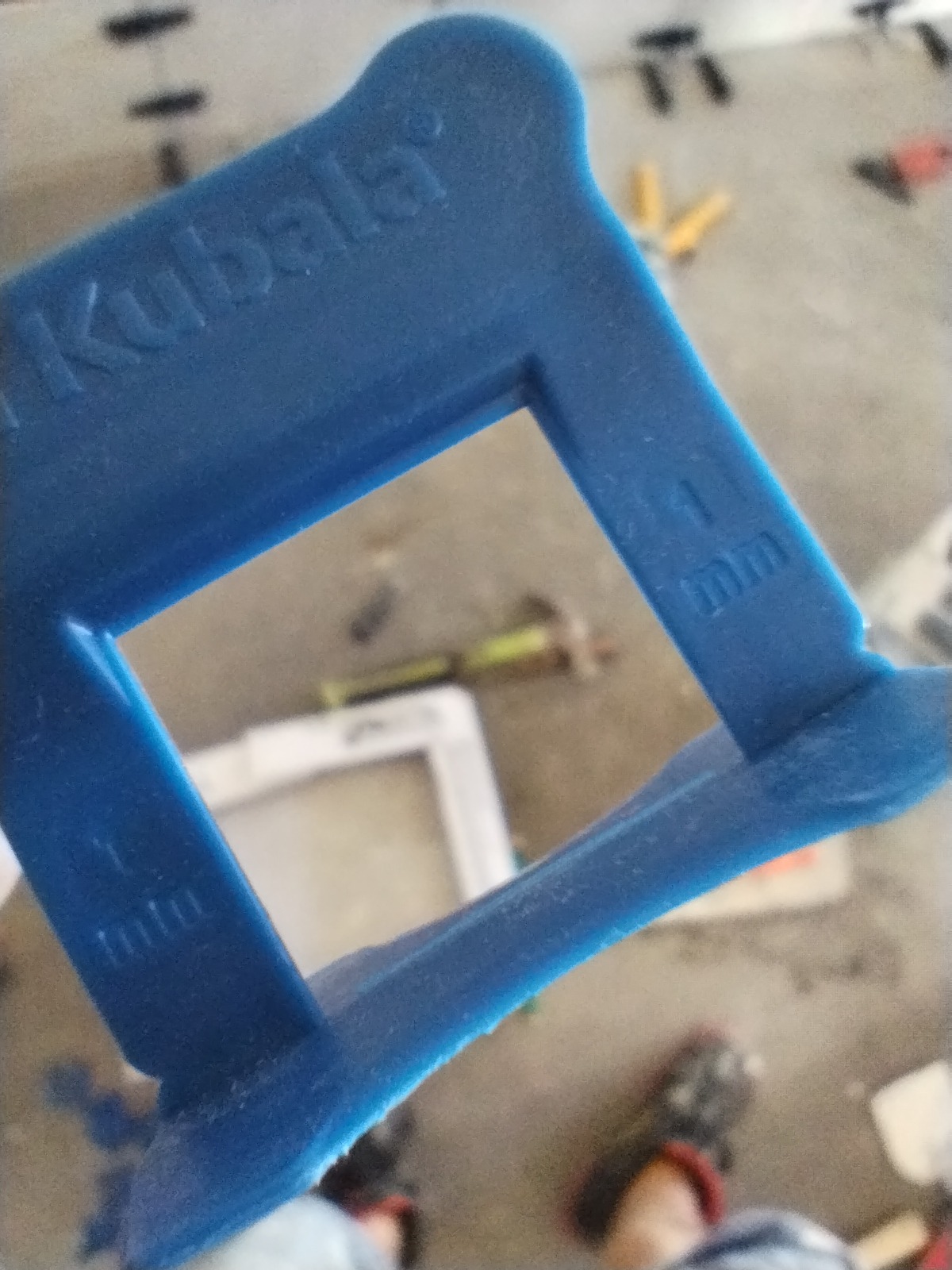 Niebieski klin dystansowy marki Kubala o grubości 1 mm, używany podczas układania płytek, widoczny na tle podłogi z resztkami zaprawy i innymi narzędziami.
