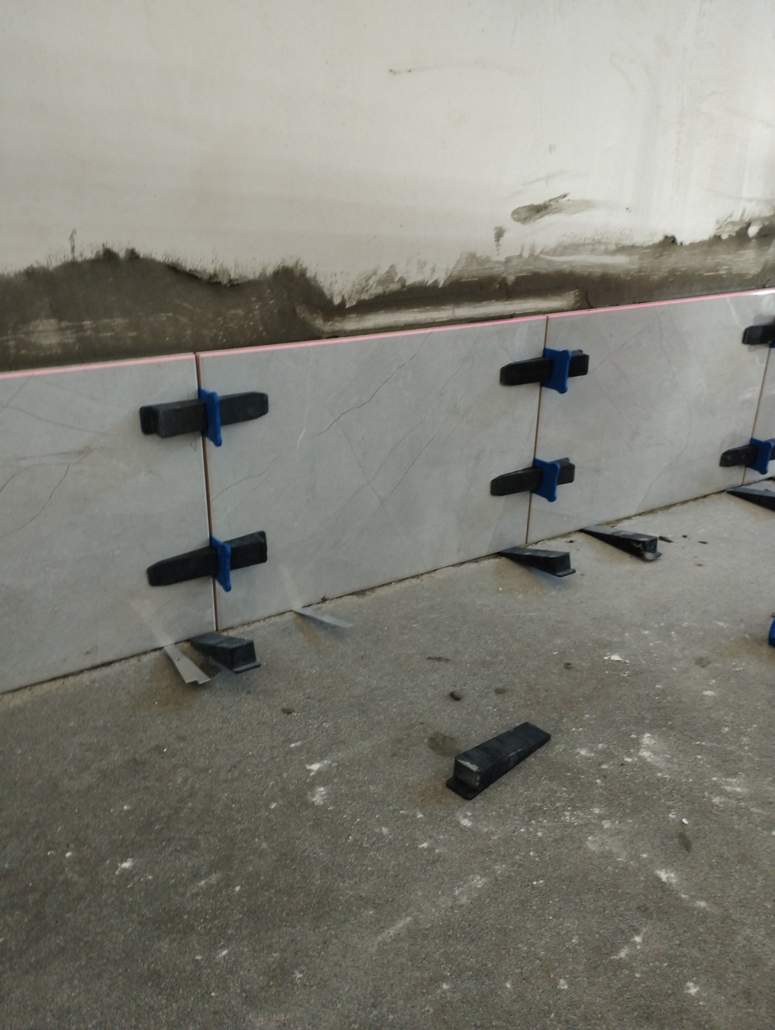 Świeżo ułożone szare płytki ścienne z systemem poziomowania, widoczne niezagruntowane ściany i surowa podłoga.