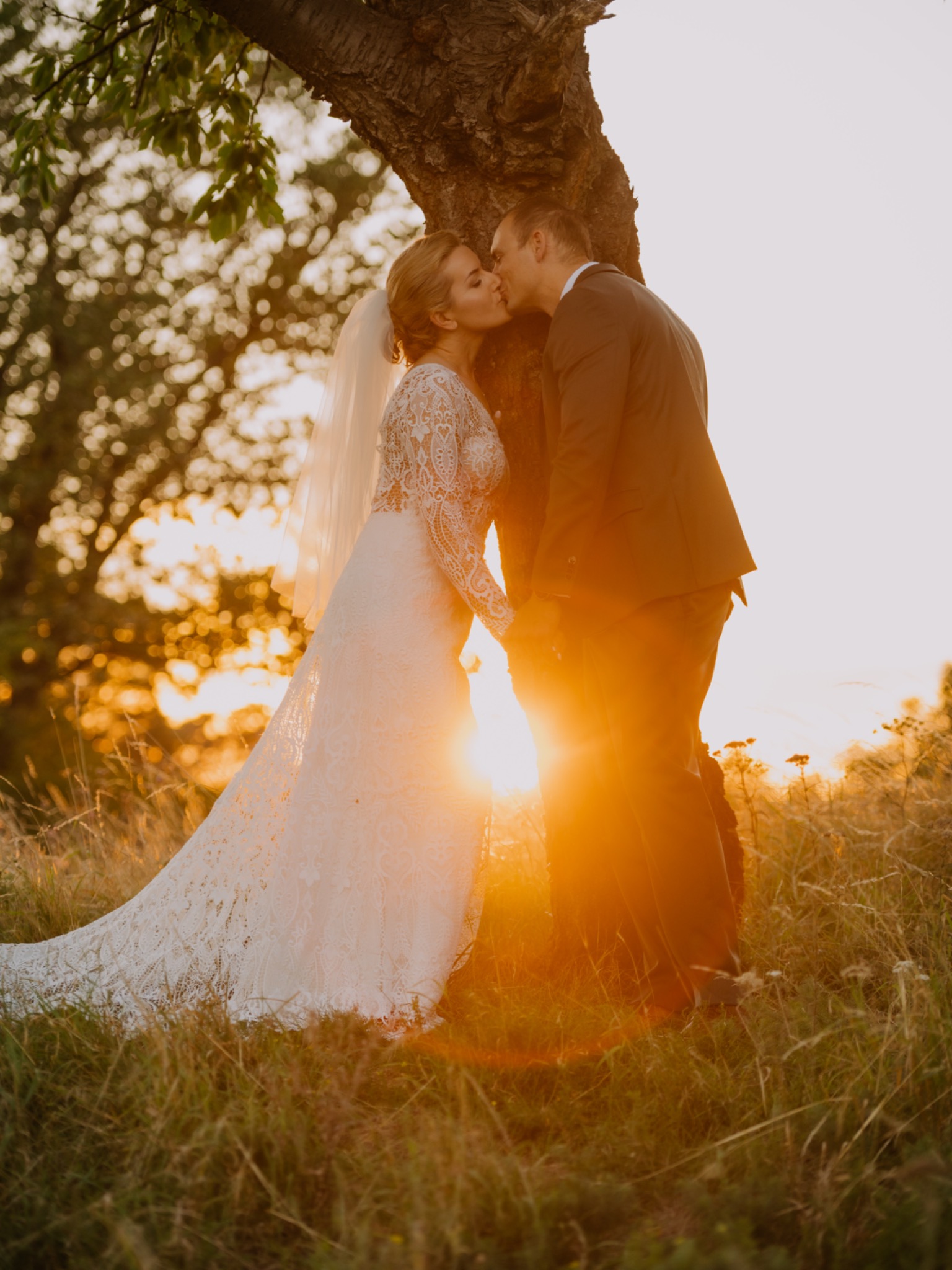 Para młoda całująca się pod drzewem w promieniach zachodzącego słońca, panna młoda w koronkowej sukni z długim rękawem i welonem, pan młody w ciemnym garniturze, naturalne tło z wysoką trawą.