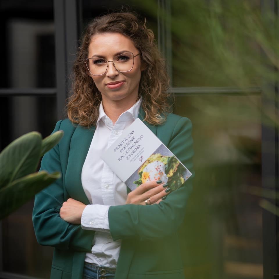 Portret kobiety w okularach, trzymającej książkę o racjonalnym żywieniu, ubrana w zieloną marynarkę na tle okna.